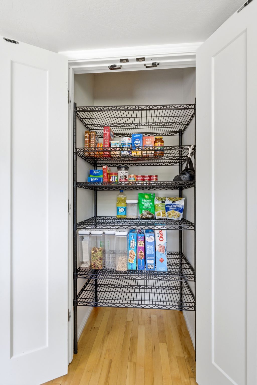 Spacious pantry featuring six tiers of black wire shelving, double white panel doors, and wood-finish flooring