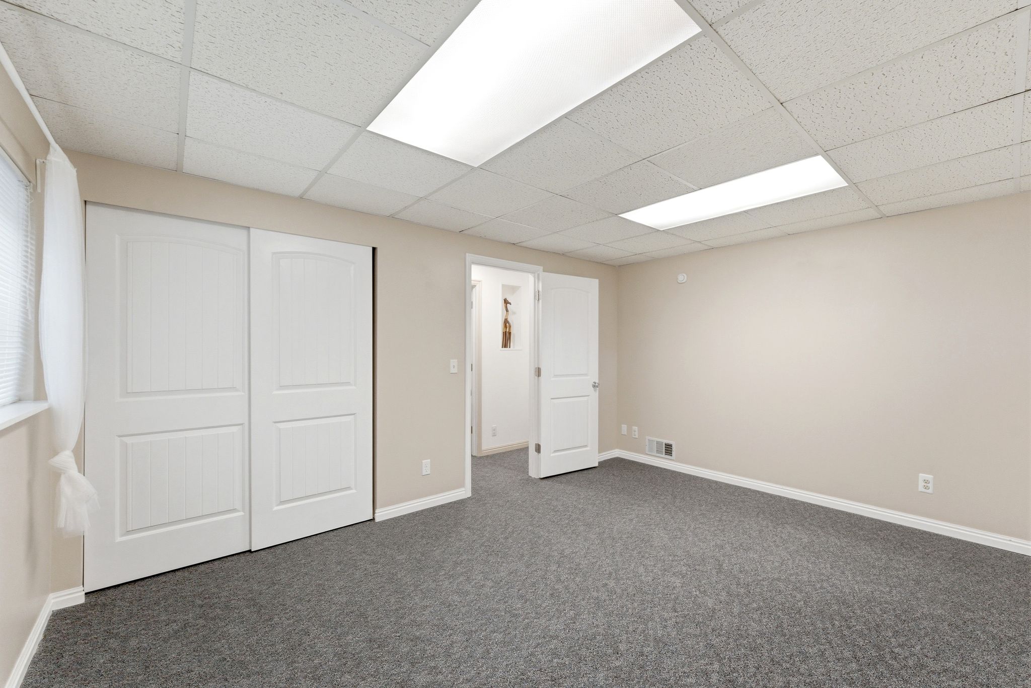 Spacious room featuring gray carpet flooring, neutral-toned walls, and a drop ceiling with integrated fluorescent lighting