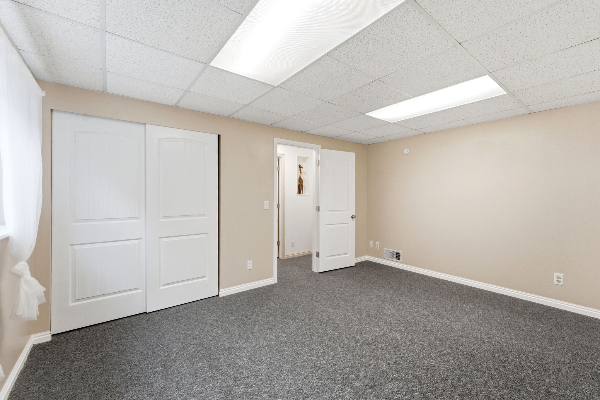 Spacious room featuring neutral carpeting, light beige walls, white baseboards, and a drop ceiling with integrated fluorescent lighting