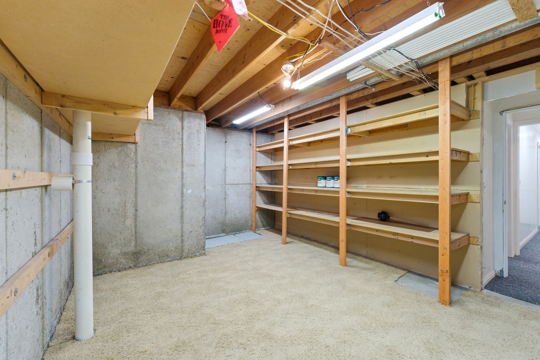 Unfinished basement area featuring concrete walls, exposed ceiling joists, built-in shelving, carpet flooring, and fluorescent lighting