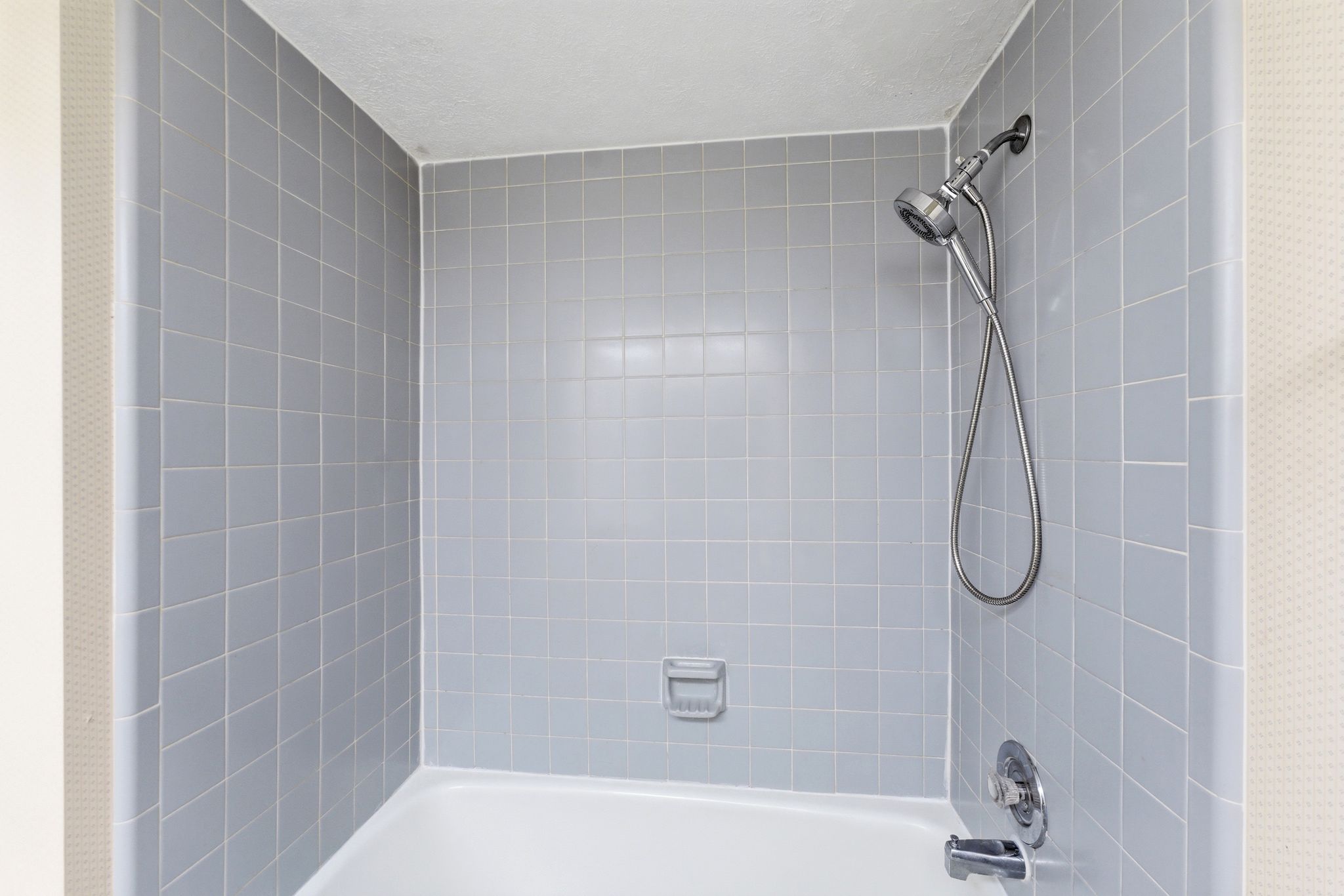 Bathtub with light gray ceramic tile surround featuring a handheld showerhead, wall-mounted faucet, and integrated soap dish