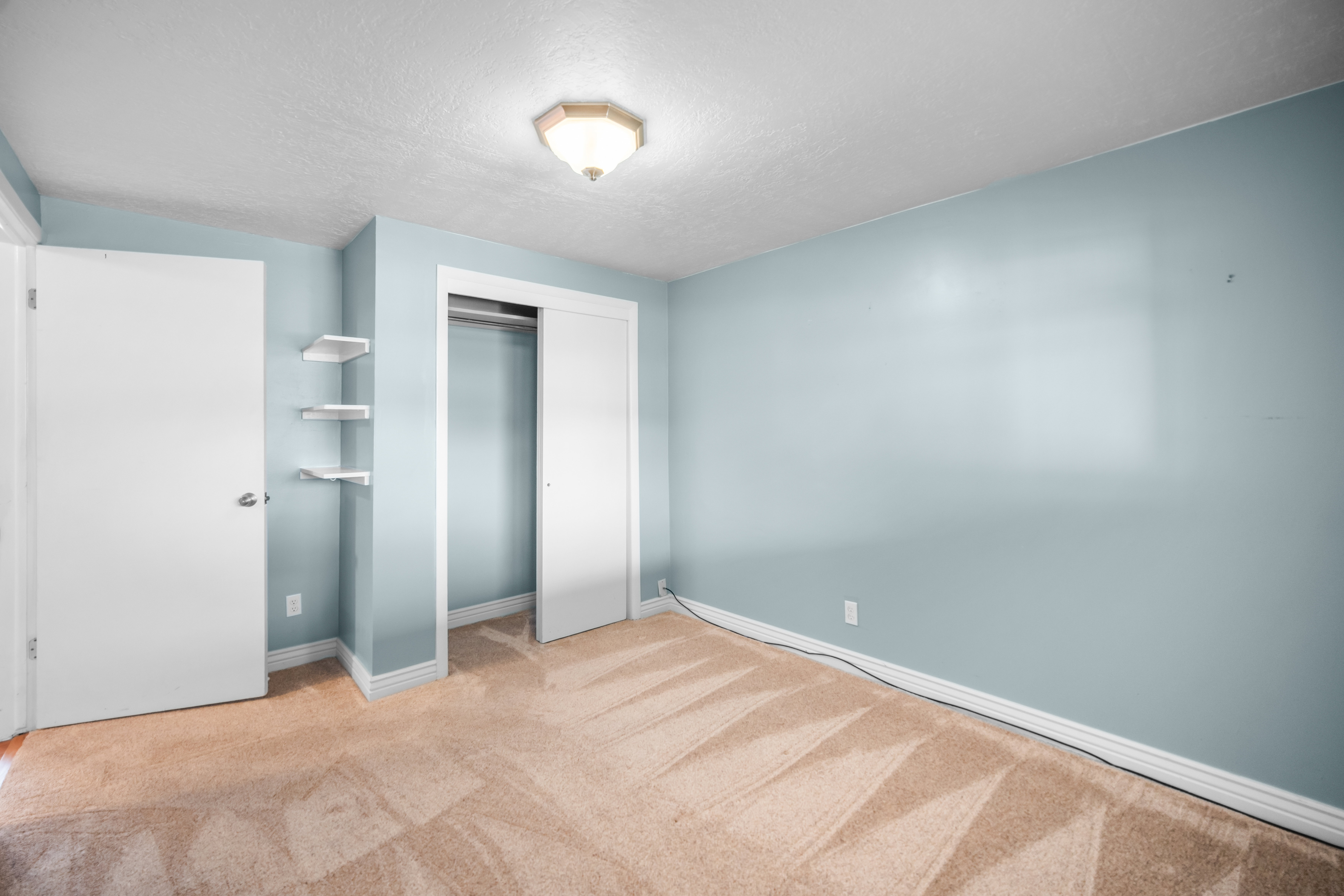 Carpeted room featuring light blue walls, white trim, a flush-mount ceiling light fixture, a closet with sliding doors, and a door with a chrome knob