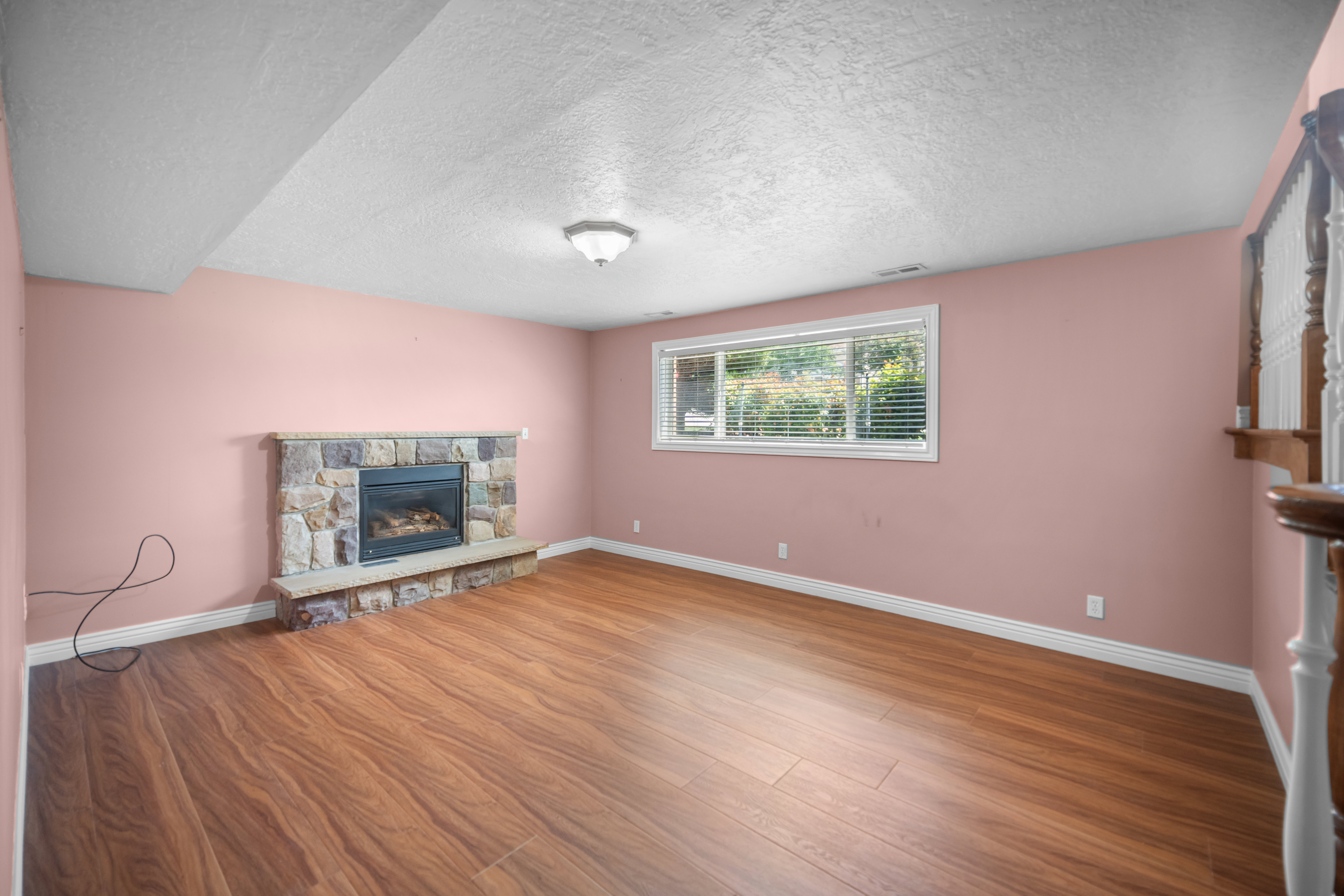 Inviting room featuring a stone-clad fireplace with hearth, a window with blinds, wood-finish flooring, and a ceiling-mounted light fixture