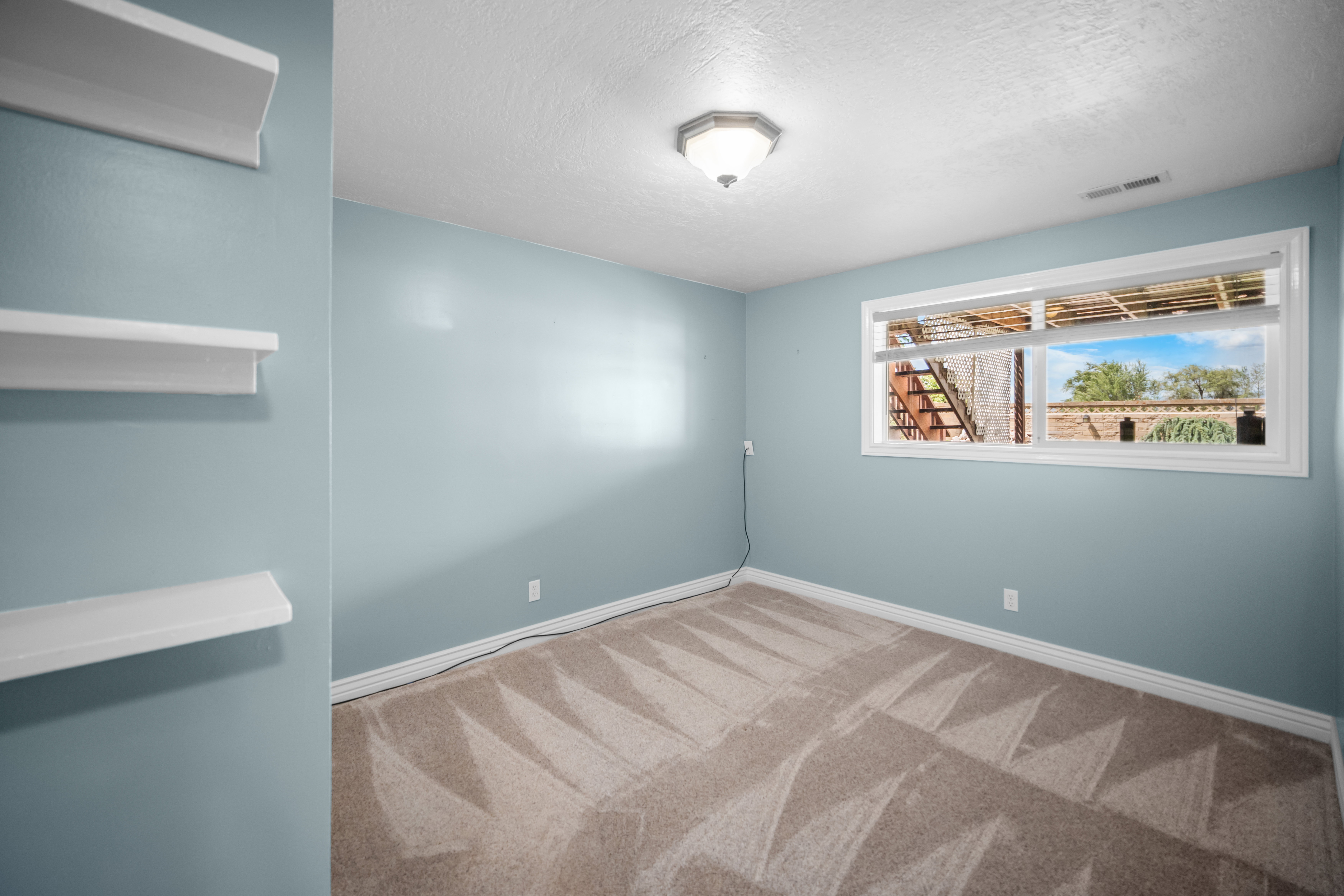 Spacious room featuring light blue walls, textured carpet flooring, and a broad window with white trim