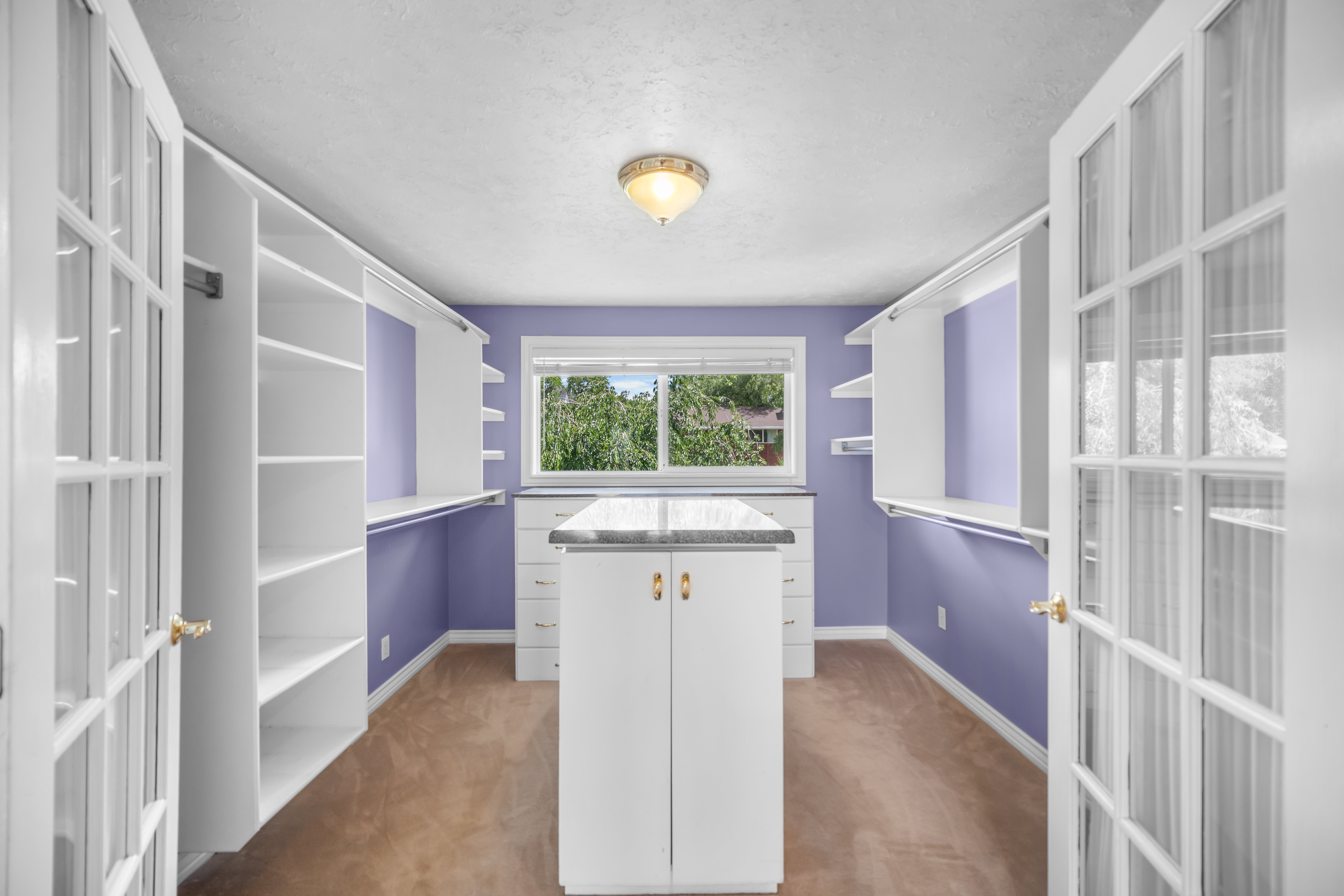 Spacious dressing room featuring a central island with a stone-finish countertop, dual French doors, built-in shelving, hanging rods, and a window for natural light