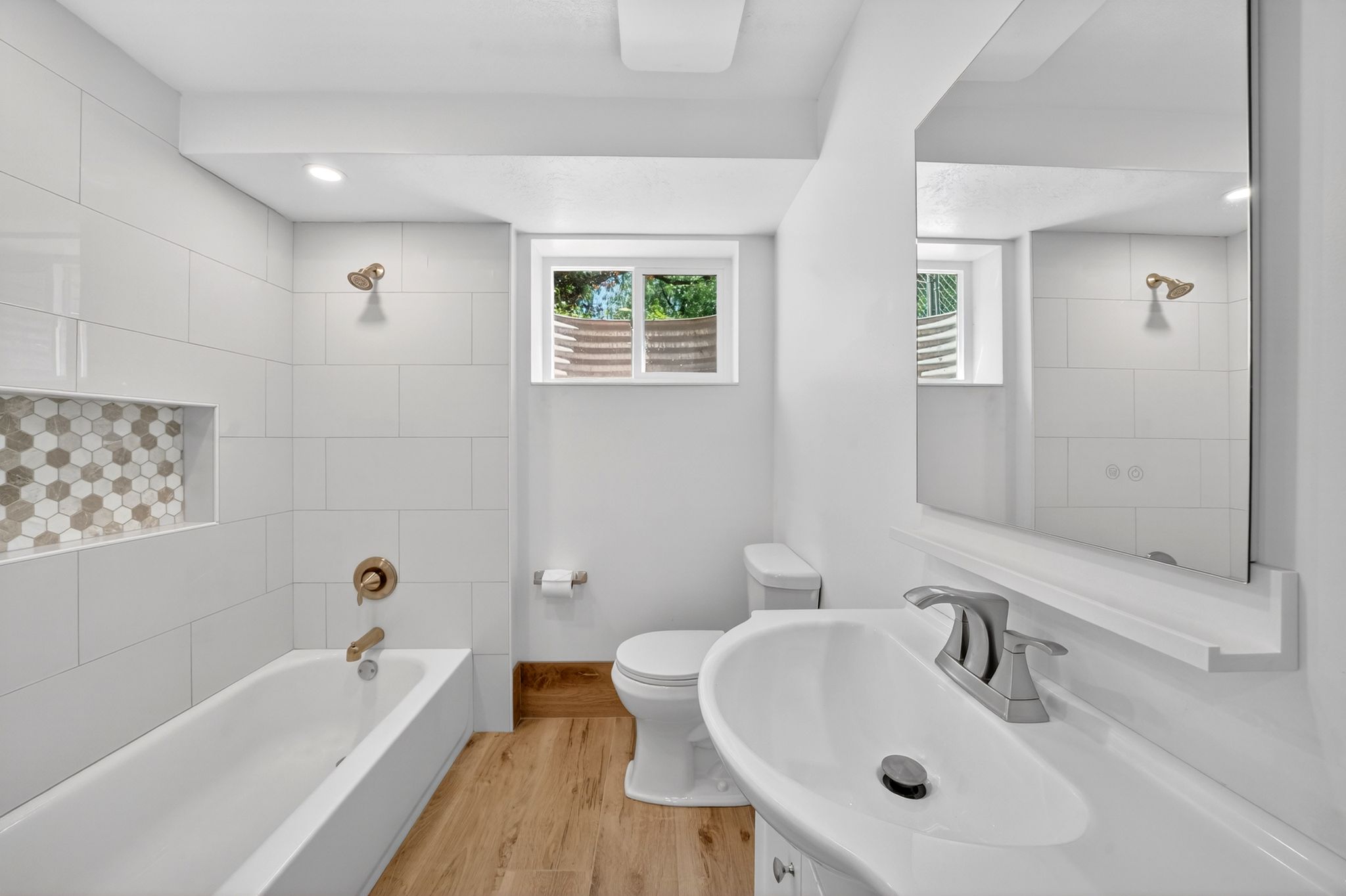 Downstairs Contemporary bathroom featuring a white tile tub surround with an integrated hexagonal mosaic niche, a deep soaking tub with brushed brass fixtures, and wood-finish flooring