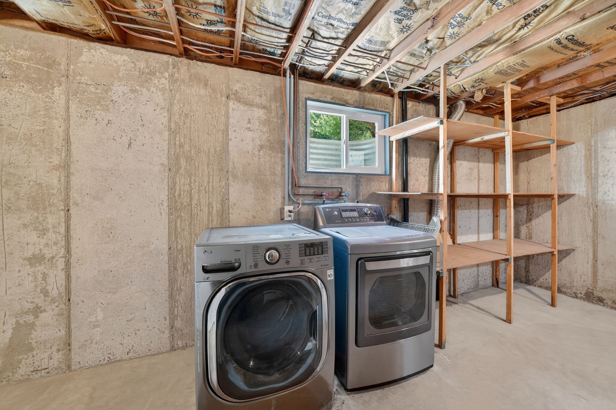 Unfinished basement space featuring exposed concrete walls, a small horizontal window, open wood shelving, and exposed ceiling joists