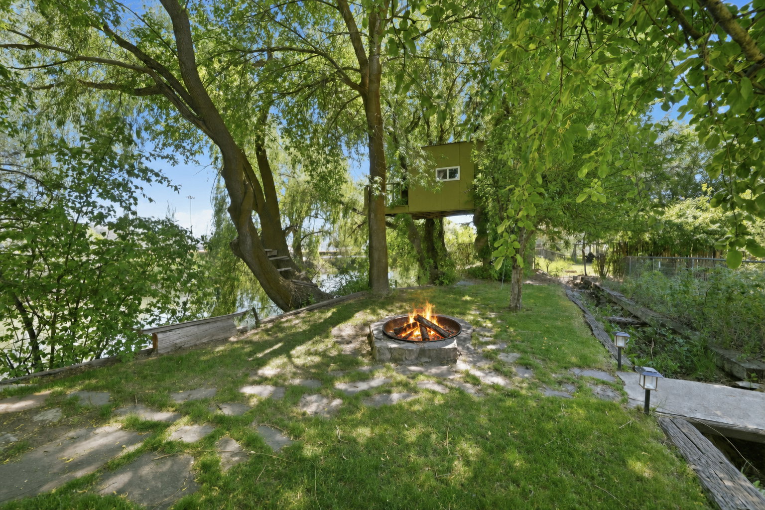 Expansive outdoor area featuring a stone fire pit, lush green lawn, mature trees, and a raised treehouse structure