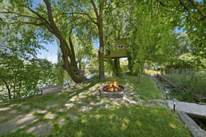 Expansive outdoor area featuring a stone fire pit, lush green lawn, mature trees, and a raised treehouse structure