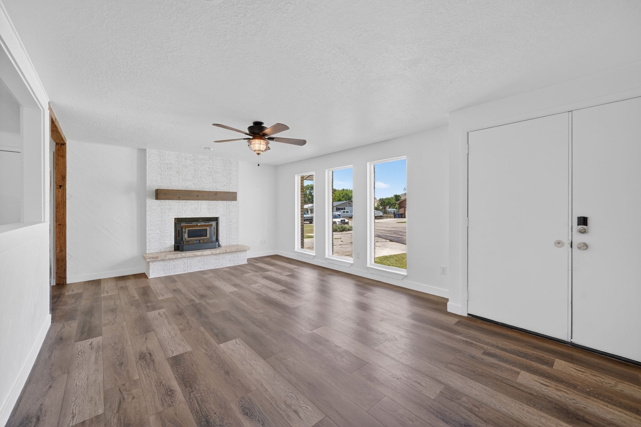 Bright living area featuring wood-finish flooring, a white textured fireplace with a stone hearth and wood mantel, three large windows, and a ceiling fan with integrated lighting