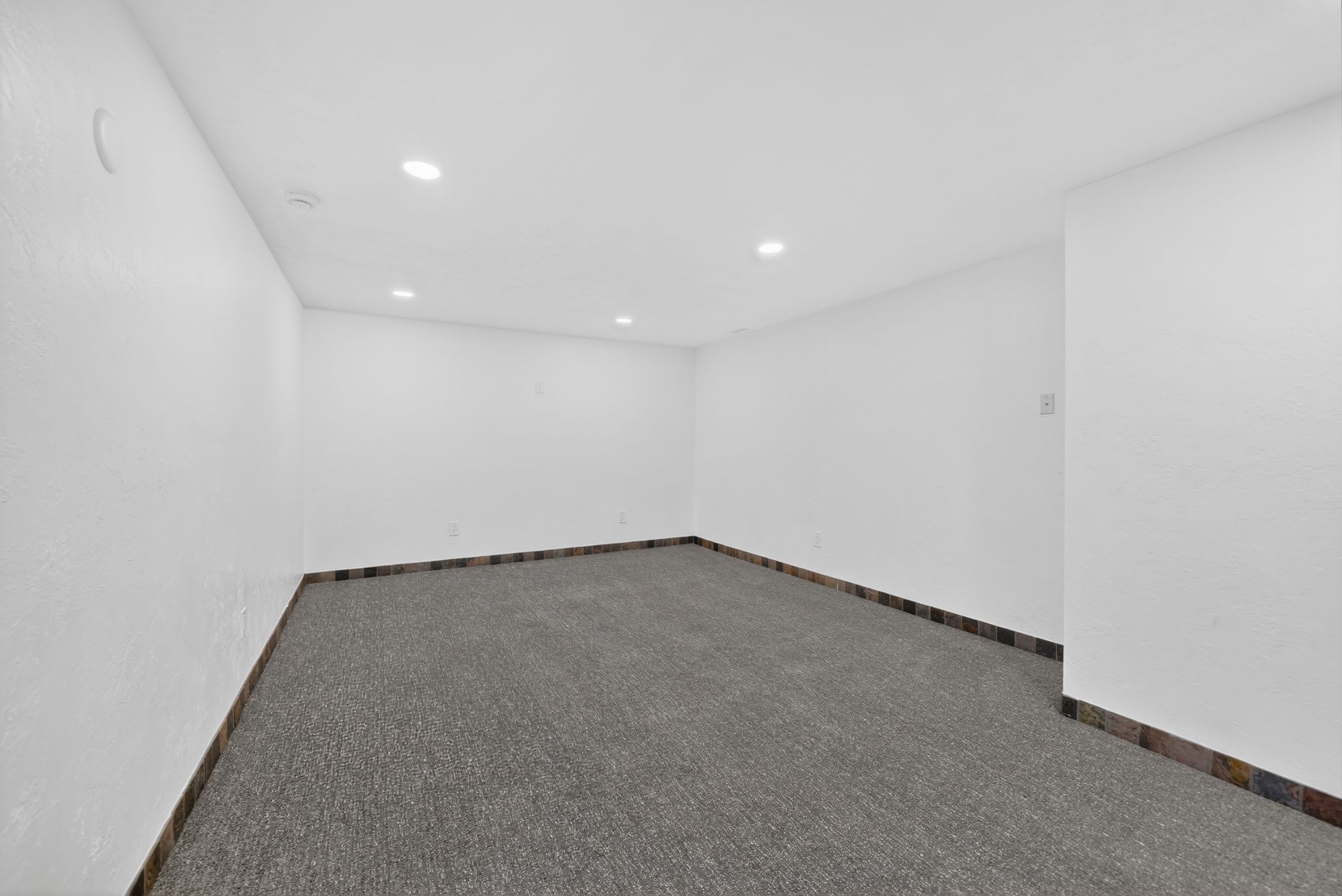 Spacious room featuring neutral carpeting, white textured walls, recessed ceiling lighting, and a dark tile baseboard