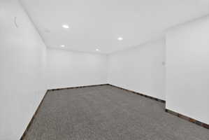 Spacious room featuring neutral carpeting, white textured walls, recessed ceiling lighting, and a dark tile baseboard