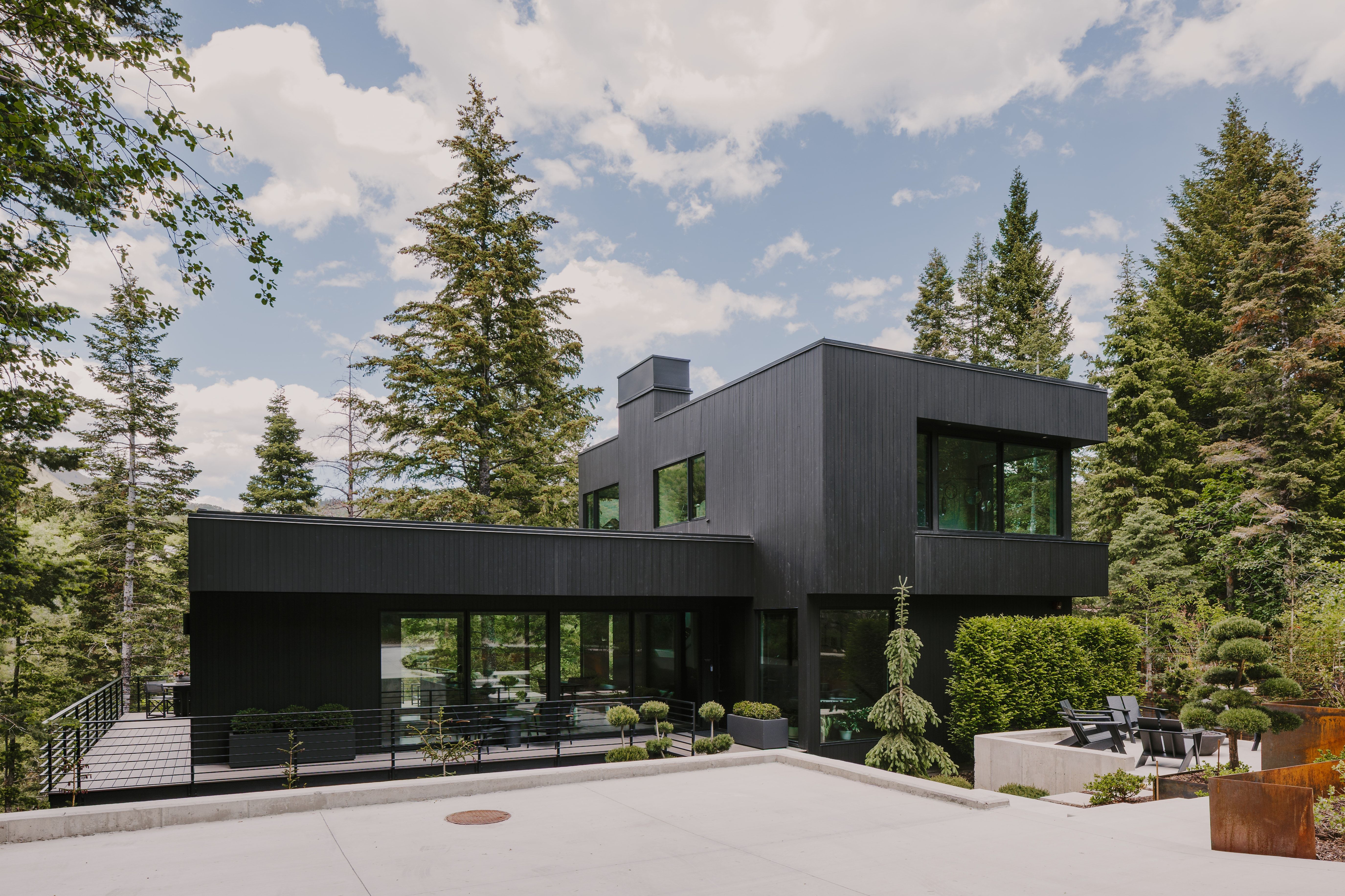 Contemporary architecture featuring a dark exterior finish, expansive windows, a concrete patio with built-in seating, a spacious outdoor deck, and mature evergreen landscaping