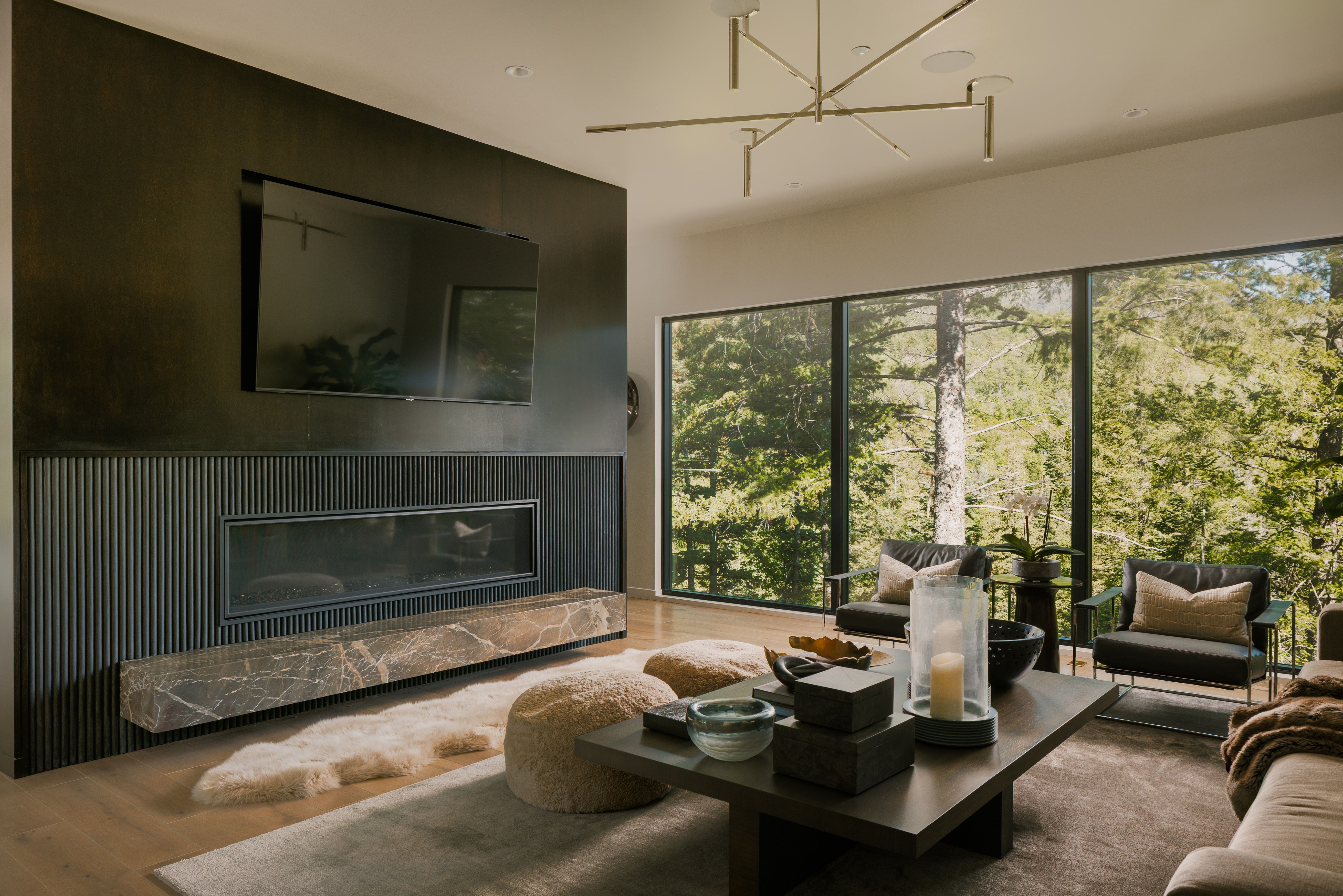 Expansive floor-to-ceiling windows with forest scenery, a linear fireplace with a floating stone hearth, wood-finish flooring, a contemporary chandelier, and a dark-toned accent wall