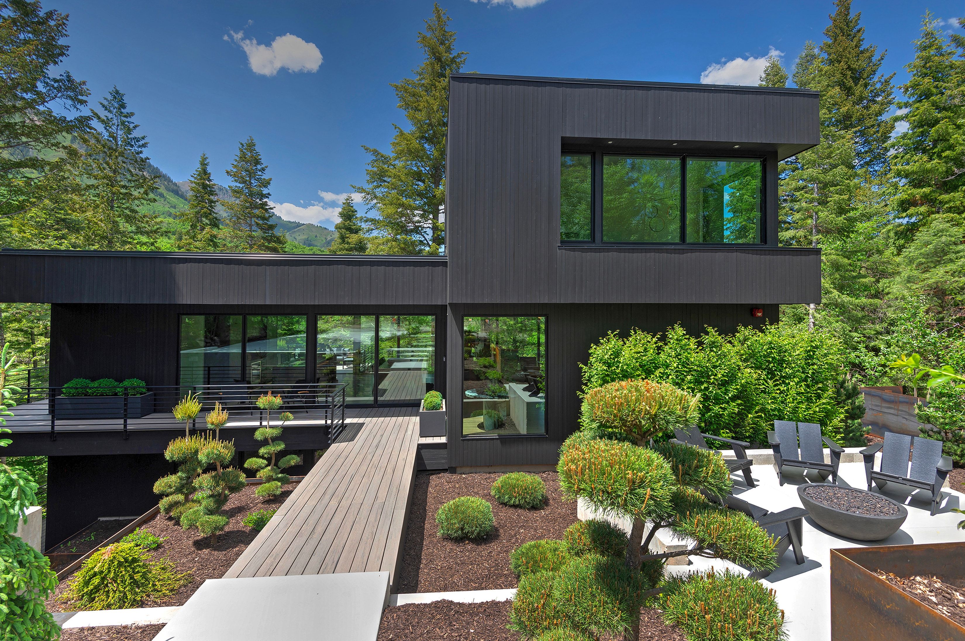 Modern black-clad exterior with extensive glazing, elevated deck, wood-finish walkway, and a concrete patio featuring a built-in fire pit
