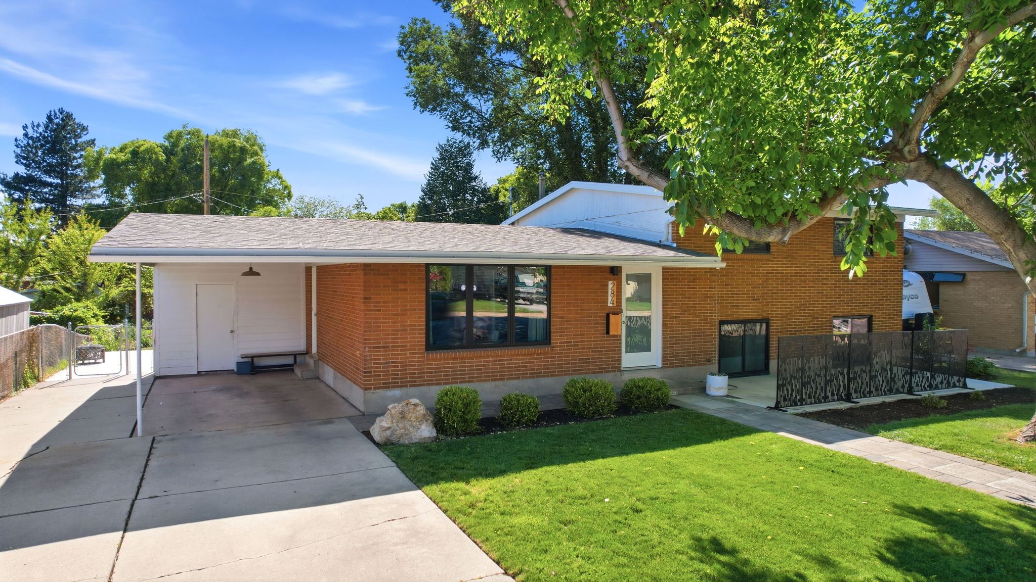 Brick exterior featuring a covered carport, mature trees, a manicured lawn, and an enclosed patio area