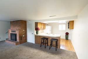 Open living area featuring a brick fireplace with hearth, an integrated kitchen counter with seating, wood-finish flooring, and light wood cabinetry