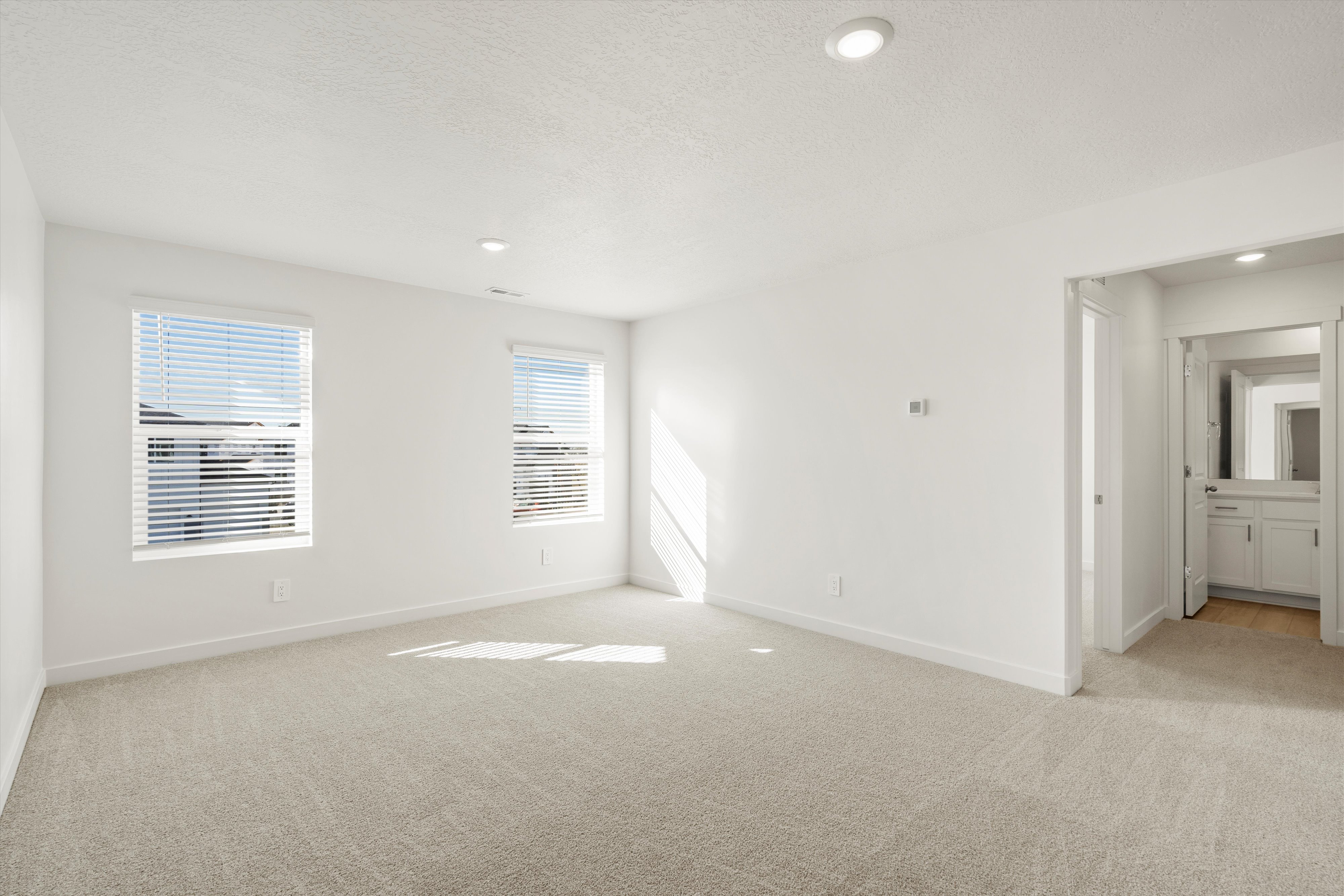 **DISCLAIMER** Interior package may vary. Carpeted room featuring white walls, two windows with blinds, recessed lighting, and a doorway opening to a bathroom with a white vanity and mirror