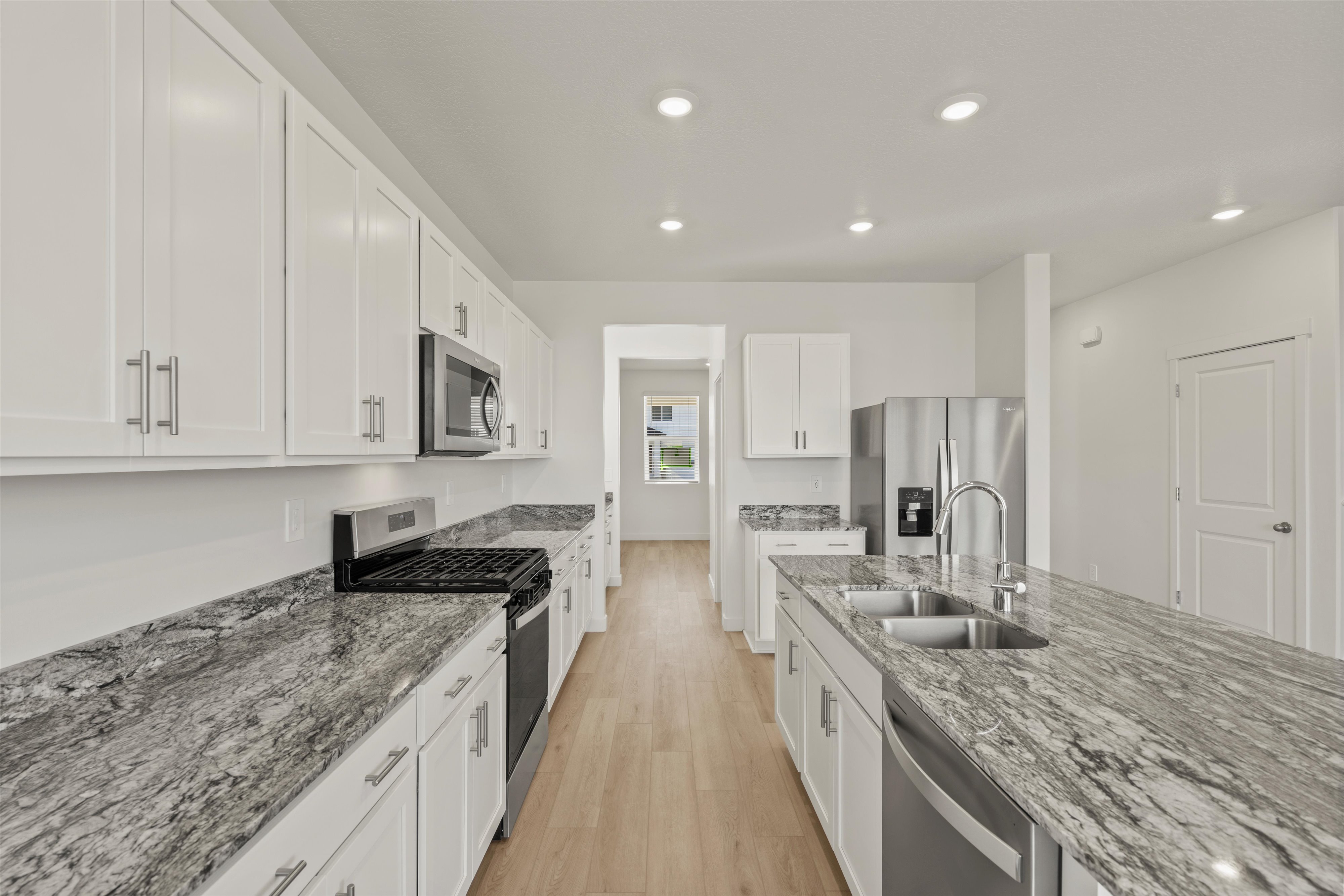 **DISCLAIMER** Interior package may vary. Kitchen featuring white shaker-style cabinetry, stainless steel appliances, speckled stone countertops, a double basin sink, and wood-finish flooring