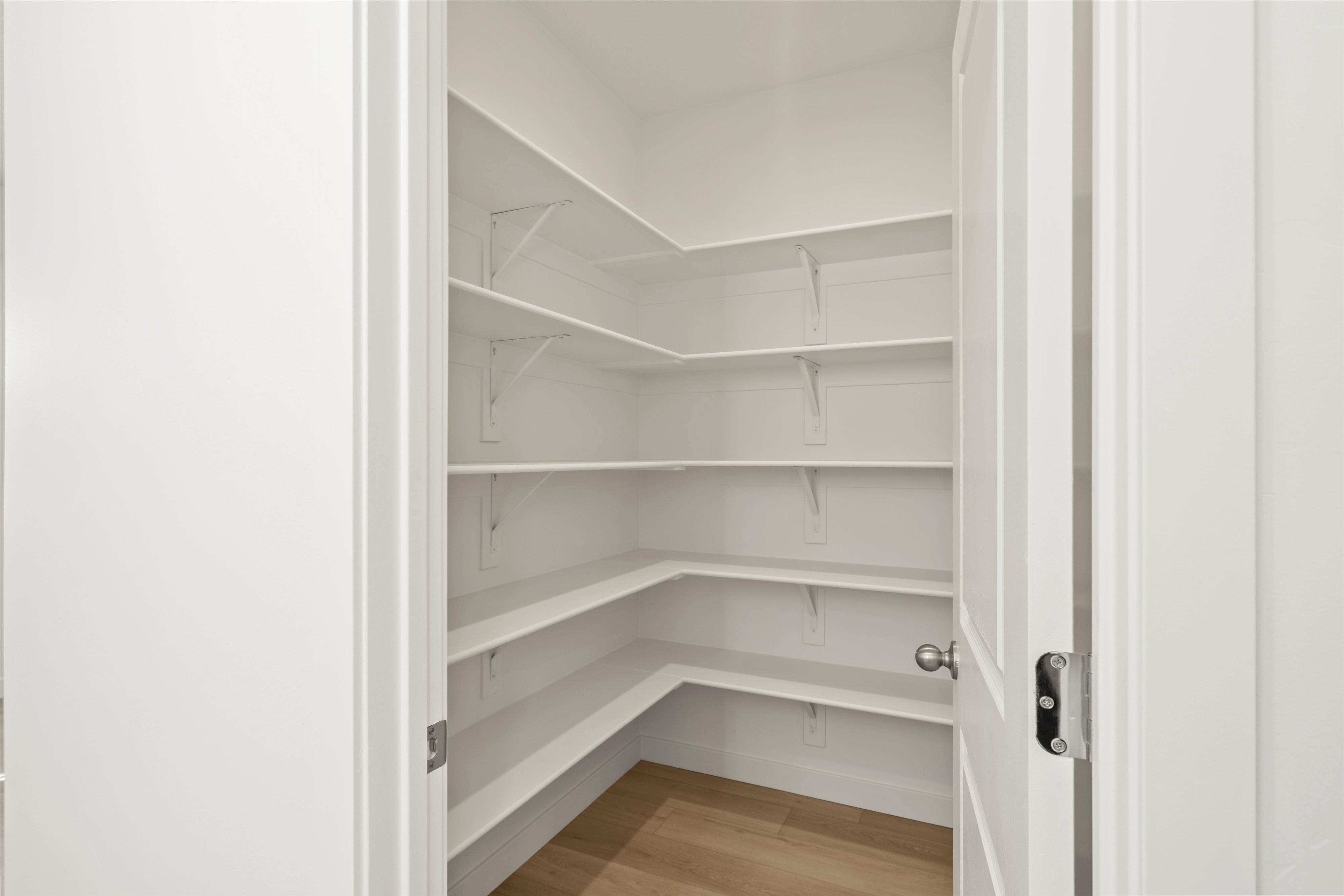 **DISCLAIMER** Interior package may vary. Spacious walk-in pantry featuring multiple levels of white shelving, wood-finish flooring, and a white interior door with a chrome knob