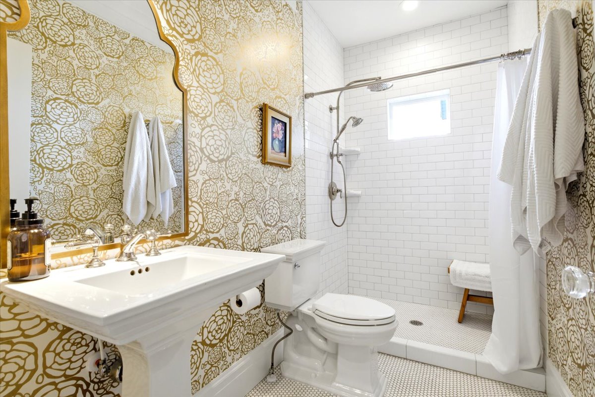 Bathroom featuring gold floral-patterned wallpaper, a white pedestal sink with polished chrome fixtures, and a shower area with white subway tile