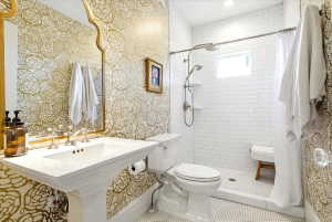 Bathroom featuring gold floral-patterned wallpaper, a white pedestal sink with polished chrome fixtures, and a shower area with white subway tile