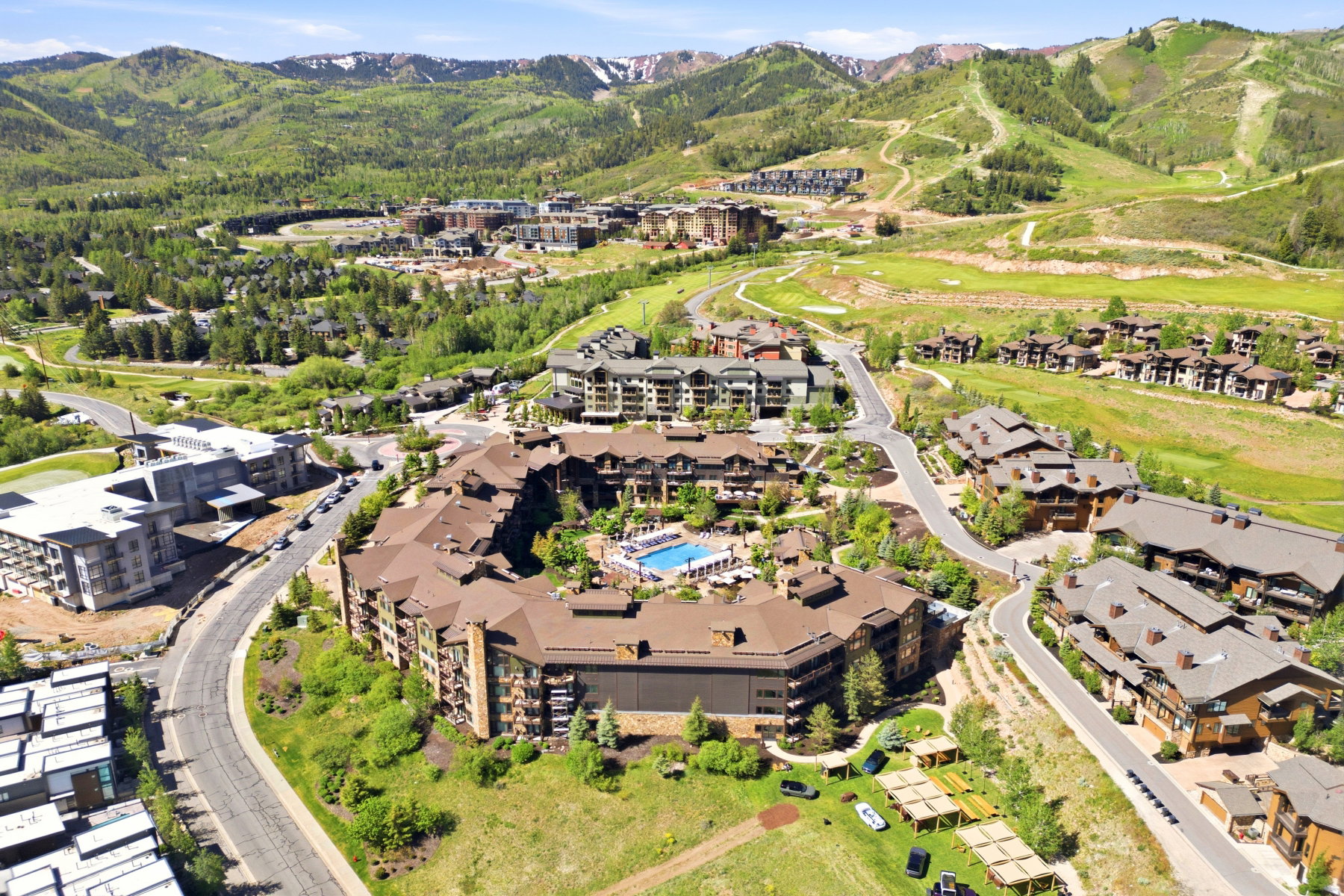 Expansive mountain development featuring a large central building with a pool, multiple surrounding residences, green landscaping, and distant ski slopes