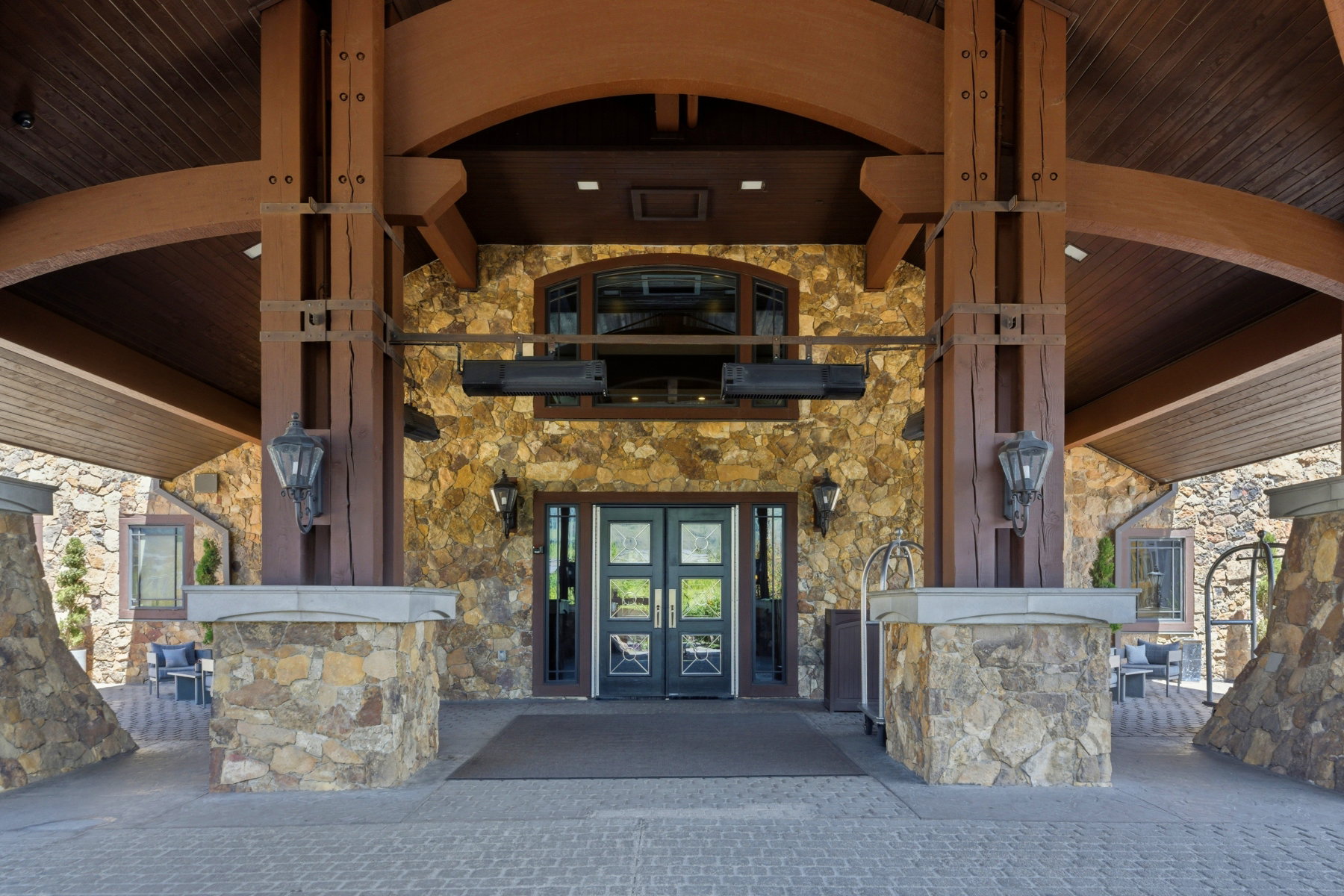 Stone and wood-beam porte-cochère entrance featuring double glass-paneled doors, arched window, and integrated outdoor lighting fixtures