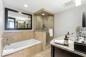 Bathroom featuring a built-in soaking tub with stone surround, a glass-enclosed shower with stone tiling, a vanity with a granite countertop and integrated sink, a large framed mirror, and recessed lighting