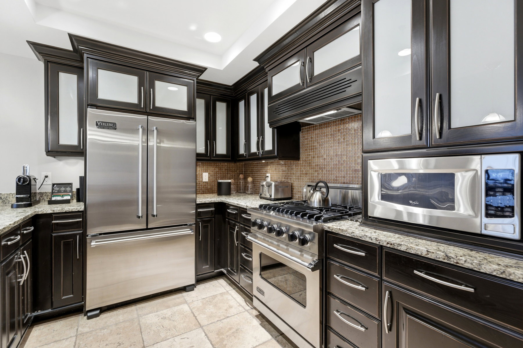 Gourmet kitchen featuring dark wood cabinetry with frosted glass inserts, granite countertops, a mosaic tile backsplash, stainless steel appliances, and recessed lighting