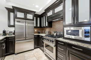 Gourmet kitchen featuring dark wood cabinetry with frosted glass inserts, granite countertops, a mosaic tile backsplash, stainless steel appliances, and recessed lighting