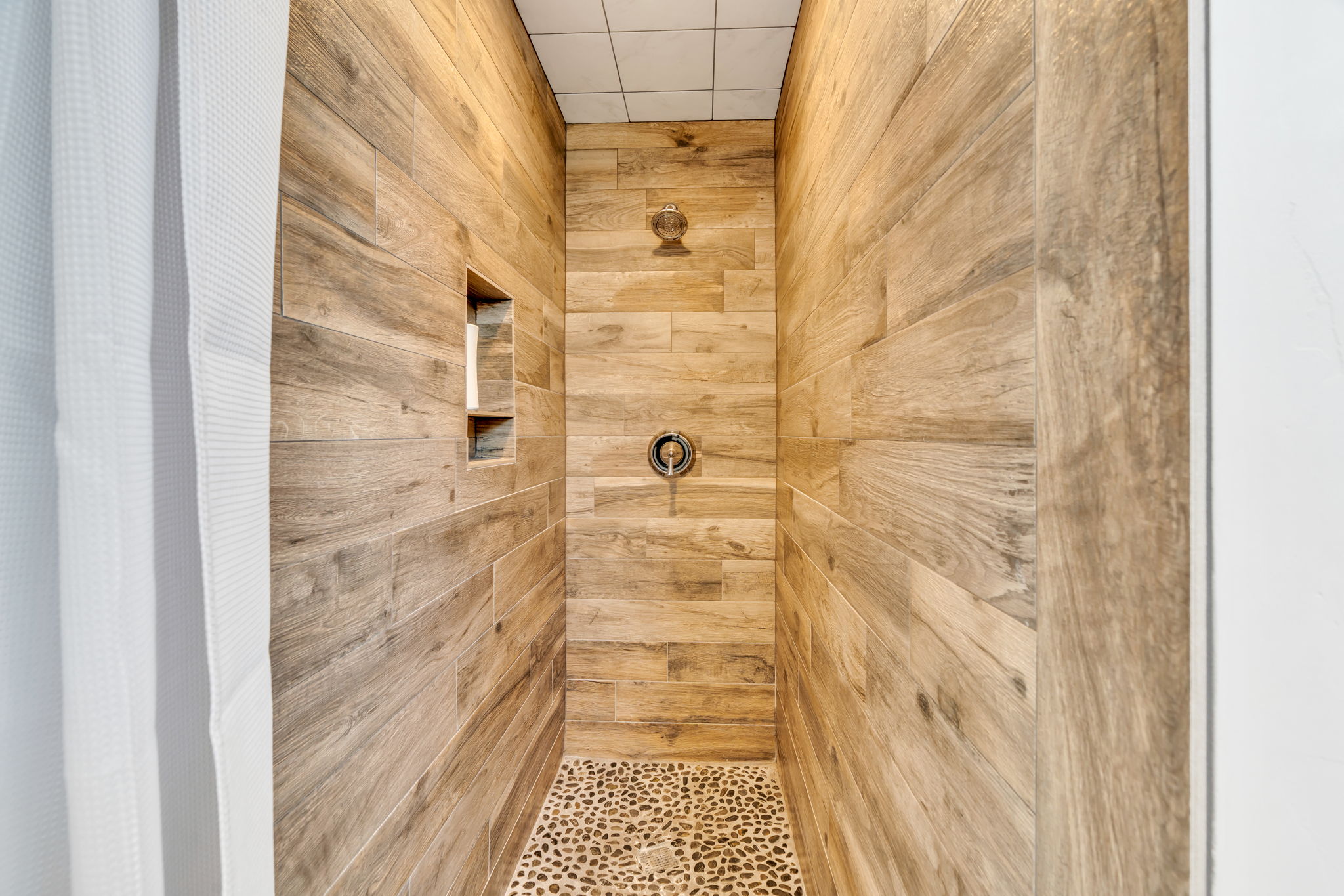 Walk-in shower featuring wood-look plank tile walls, a built-in shower niche, and a pebble tile floor