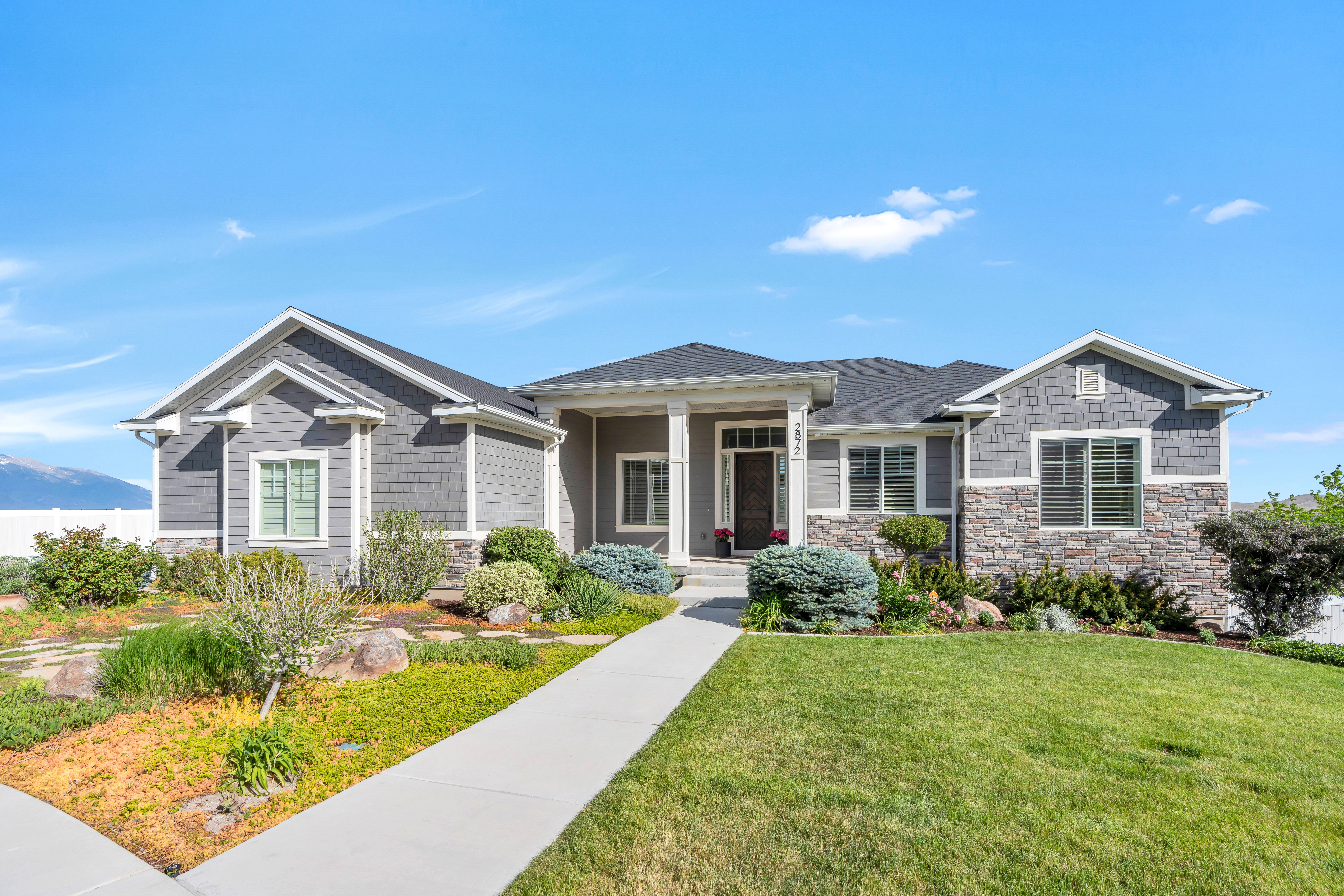 Ranch-style residence featuring a gray siding and stone facade, a covered entry with white columns, a dark shingle roof, and a mature front yard with a concrete walkway