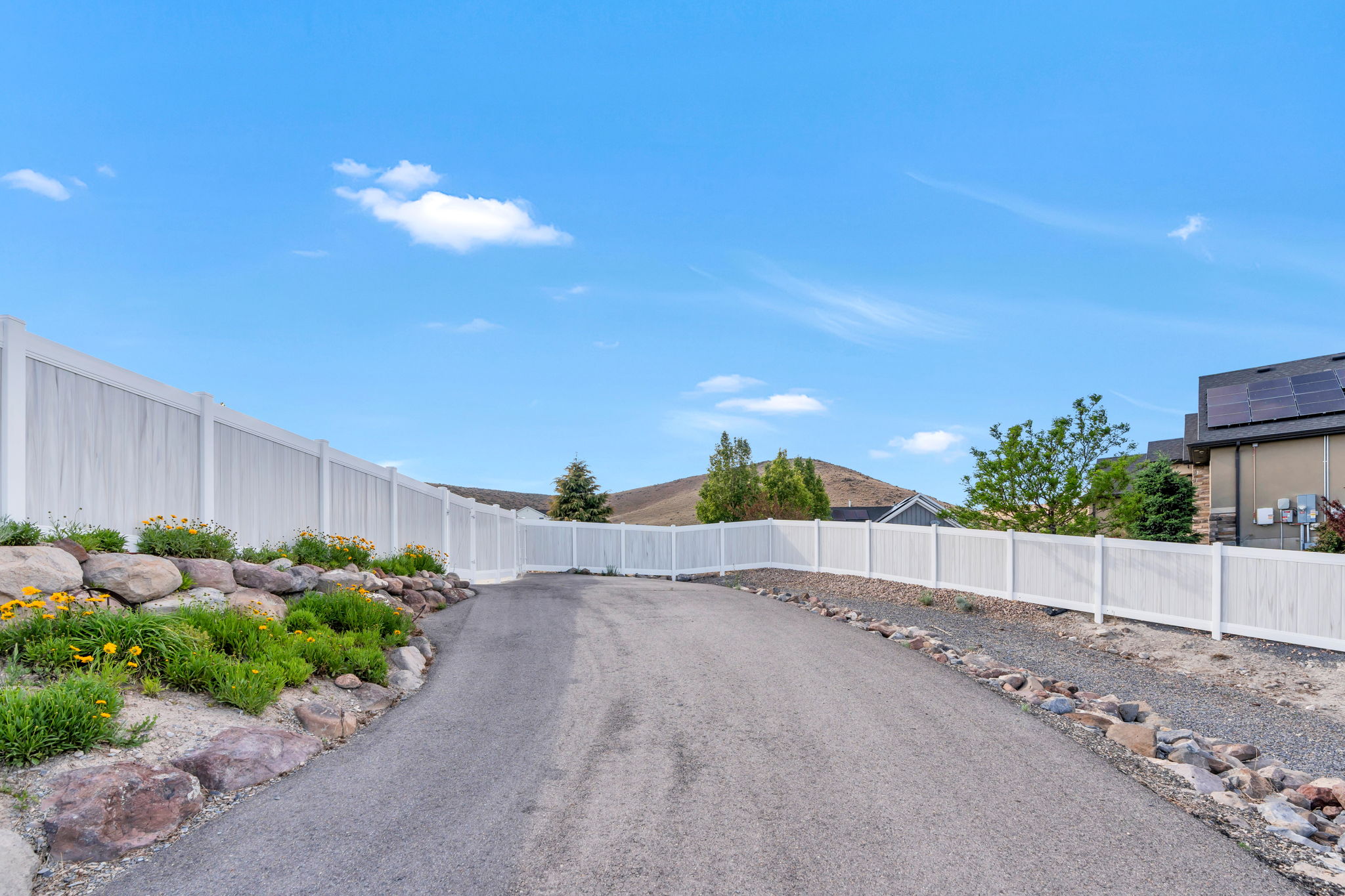 Paved driveway bordered by a white privacy fence and a rock garden with plantings
