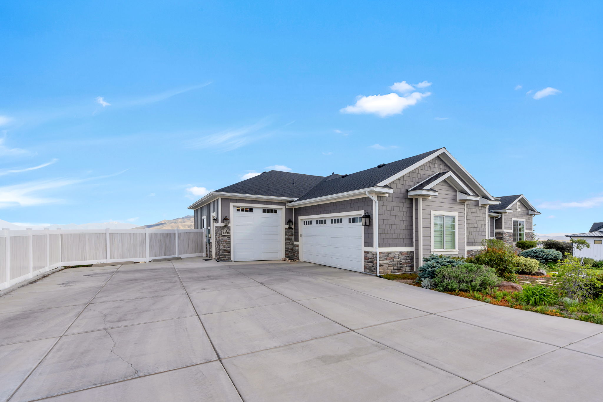 Exterior featuring gray siding, stone accents, two white garage doors, a concrete driveway, and established landscaping