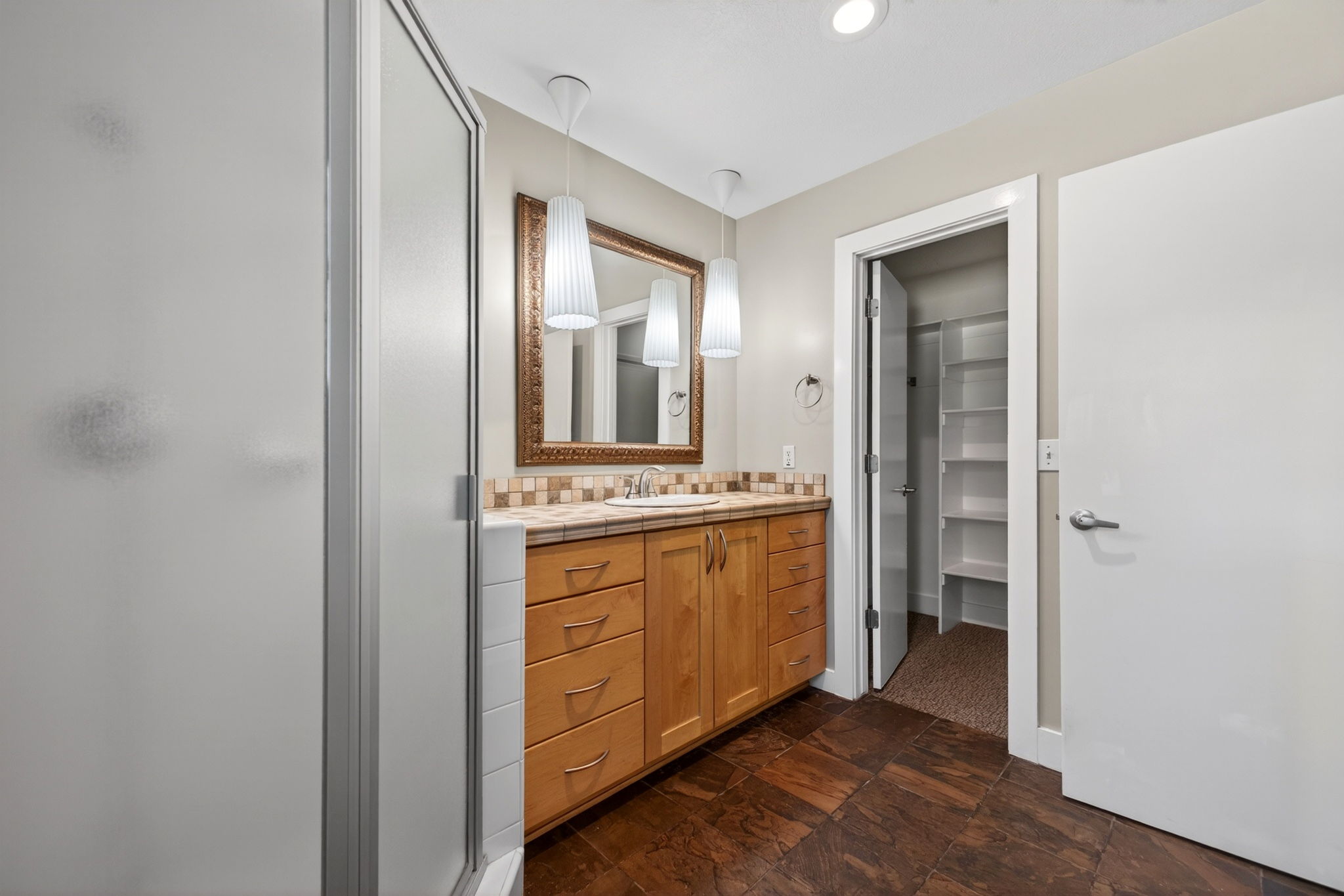 Large main bathroom featuring a spacious wood vanity, attached walk in closet, frosted glass shower, and slate tile flooring