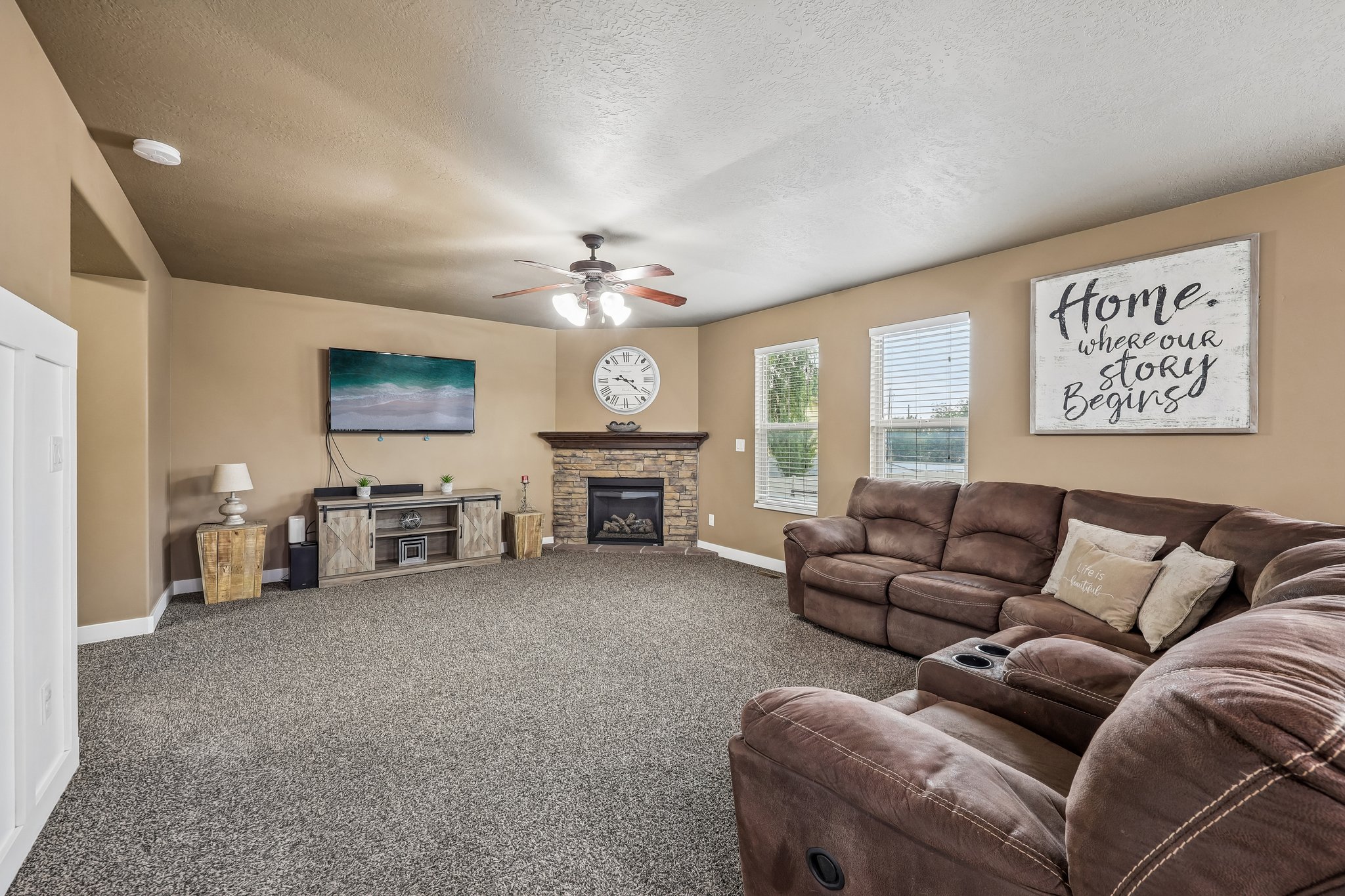 Spacious living area featuring a corner fireplace with stone surround and wood mantel, neutral carpet flooring, a ceiling fan, and multiple windows with blinds