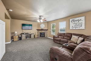 Spacious living area featuring a corner fireplace with stone surround and wood mantel, neutral carpet flooring, a ceiling fan, and multiple windows with blinds