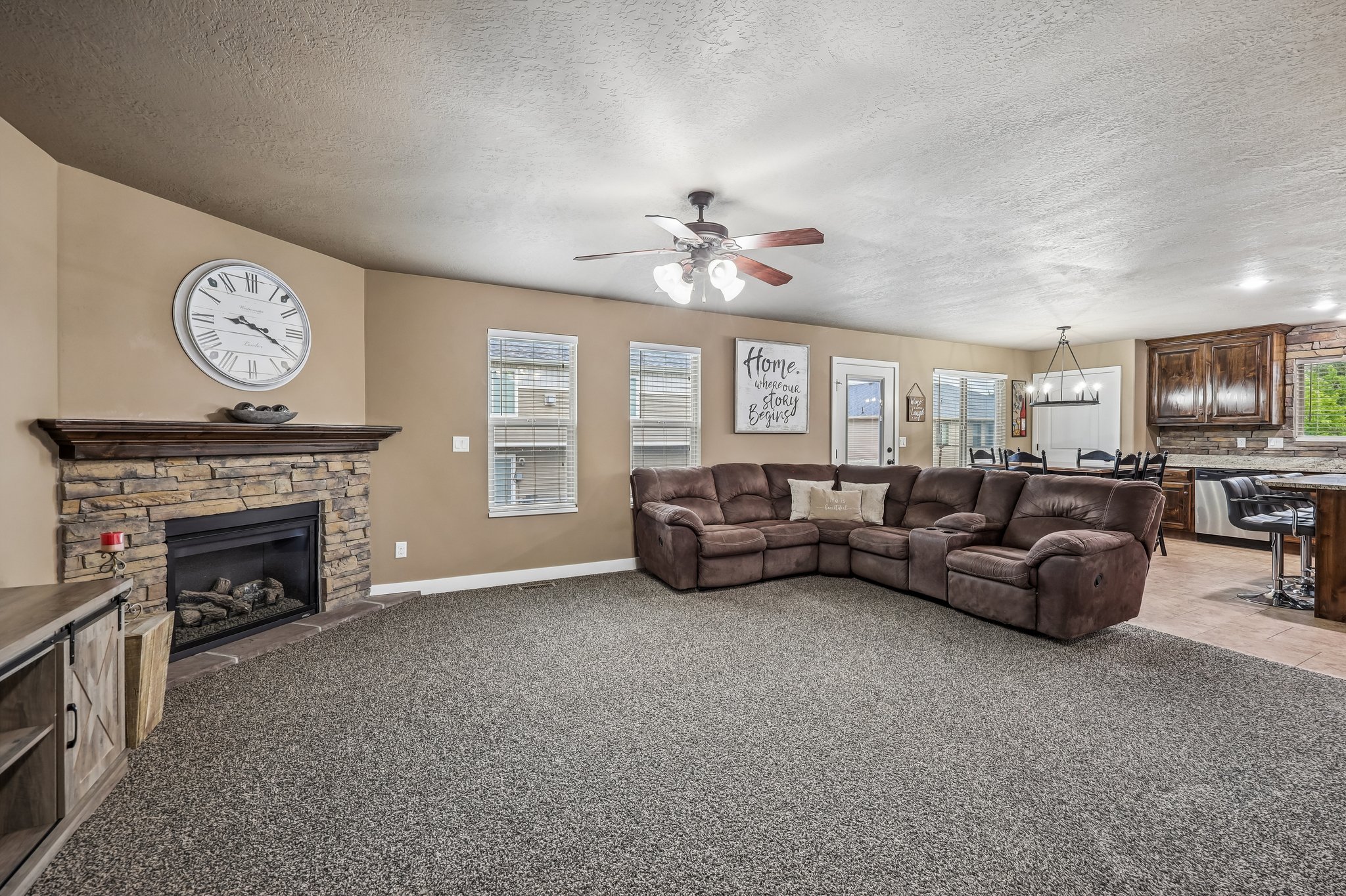 Spacious living area featuring a stone-clad fireplace with a wood mantel, carpet flooring, a ceiling fan, and multiple windows with blinds