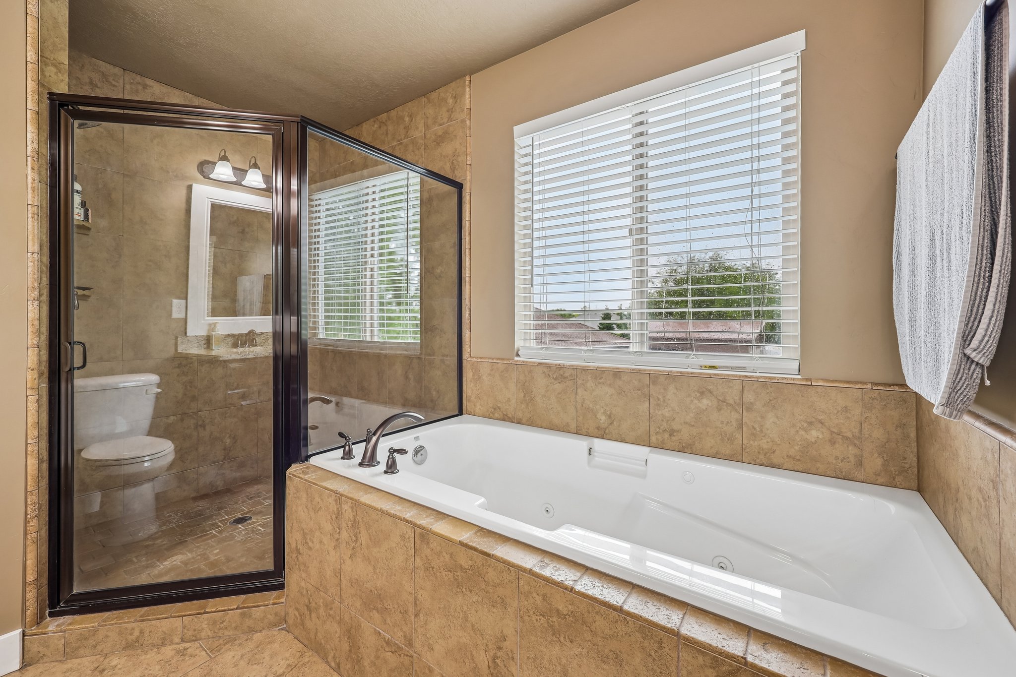 Tiled bathroom featuring a built-in corner bathtub, a glass-enclosed shower, a window with horizontal blinds, and a mounted vanity mirror