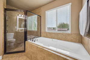 Tiled bathroom featuring a built-in corner bathtub, a glass-enclosed shower, a window with horizontal blinds, and a mounted vanity mirror