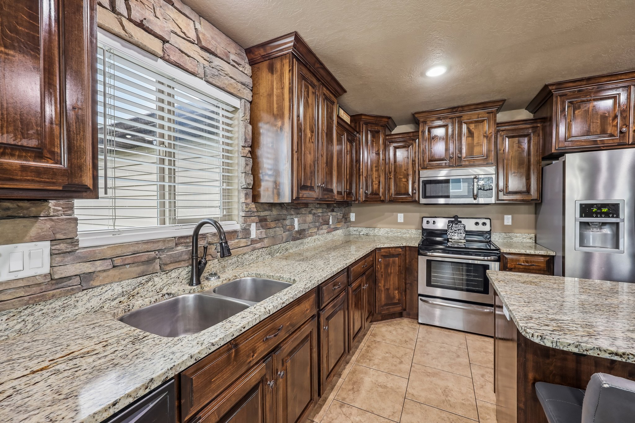 Kitchen featuring rich wood cabinetry, stone veneer backsplash, light granite countertops, and stainless steel appliances