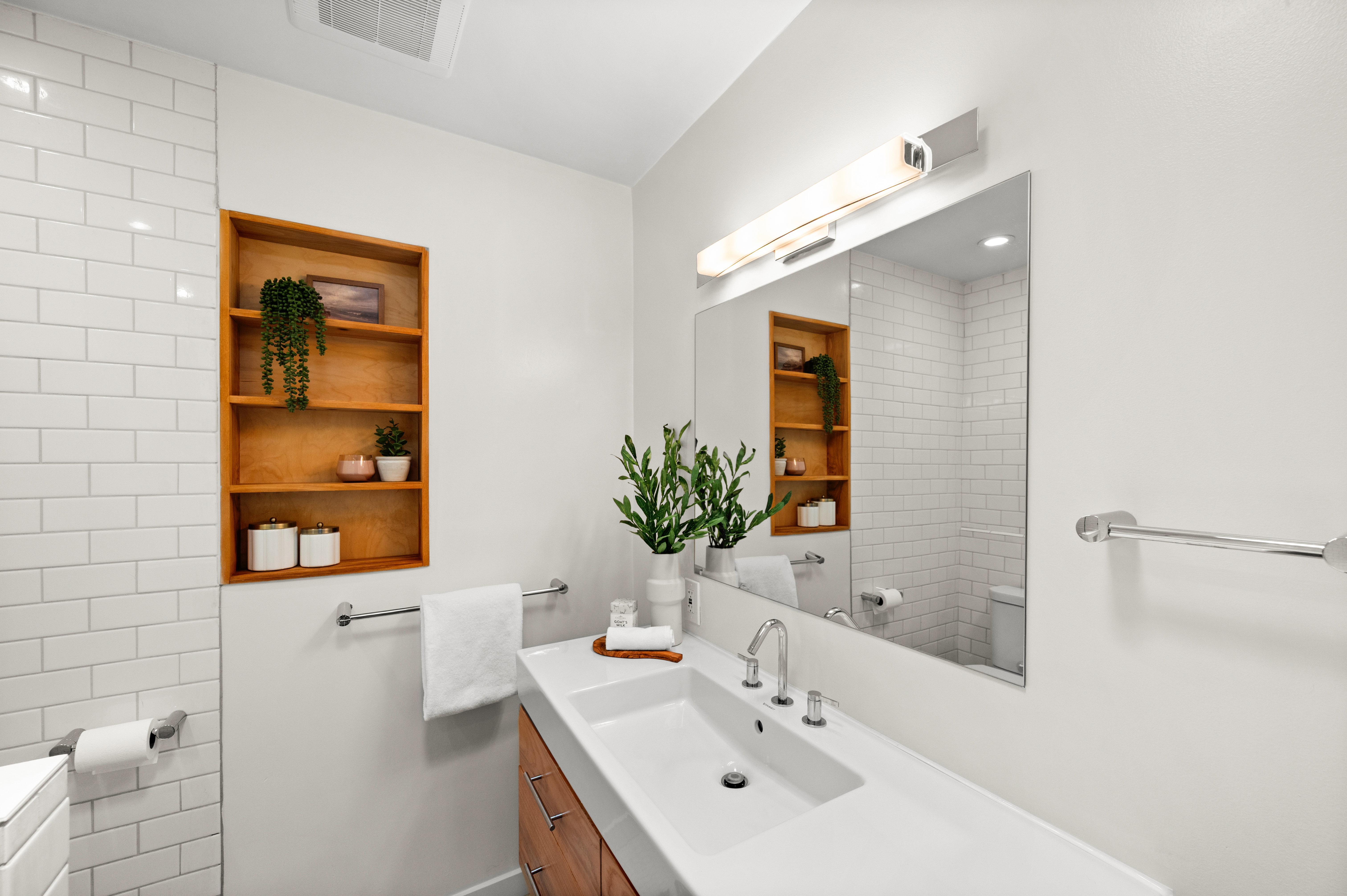 Contemporary bathroom featuring a wide integrated sink, a rectangular mirror with overhead lighting, a built-in wooden shelving unit, and subway tile accents