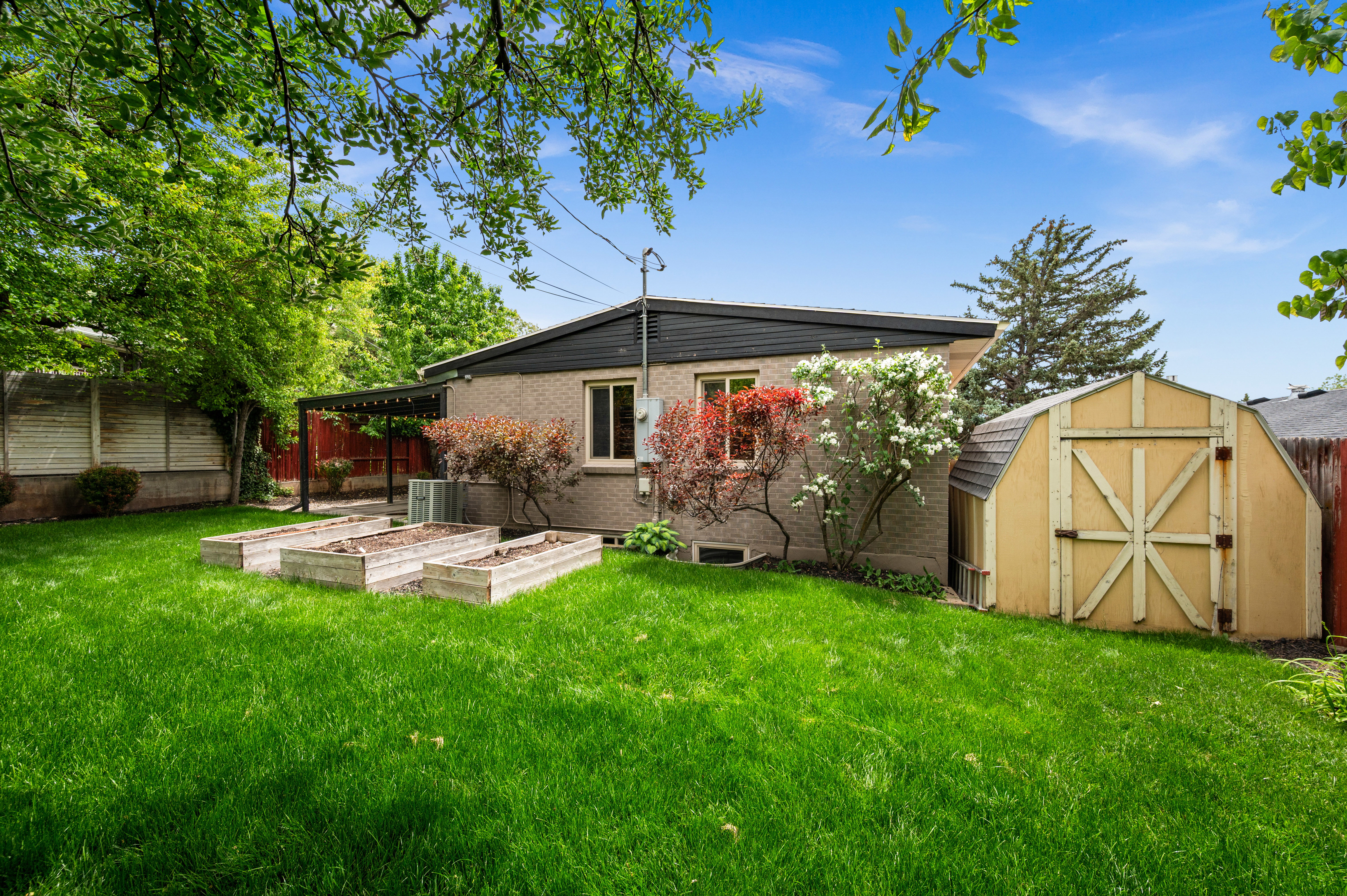 Expansive green lawn featuring three raised garden beds, a storage shed, and a covered patio area