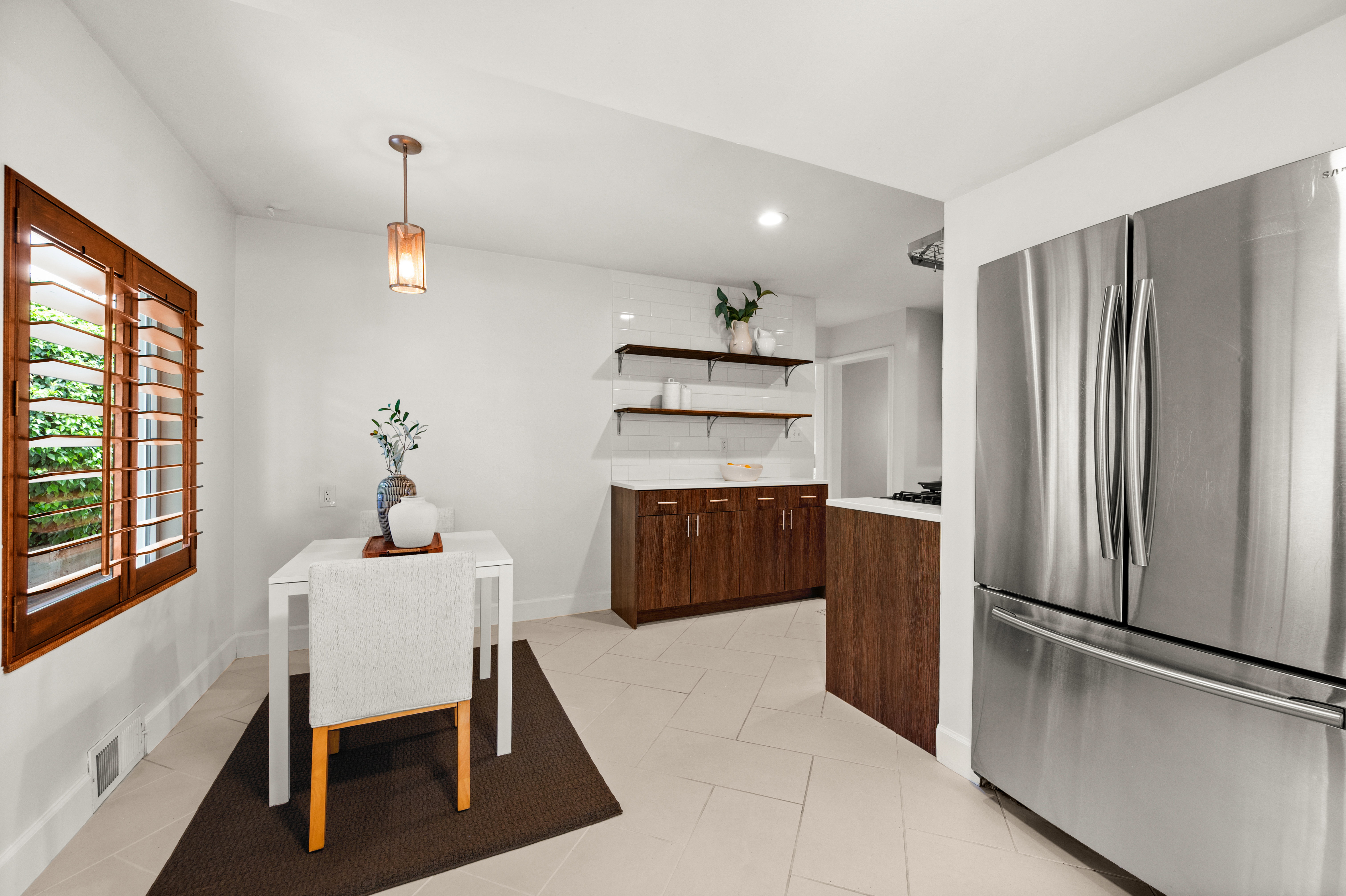 Kitchen area featuring stainless steel French-door refrigerator, light-colored tile flooring, and a dining nook with a wood-framed window and plantation shutters