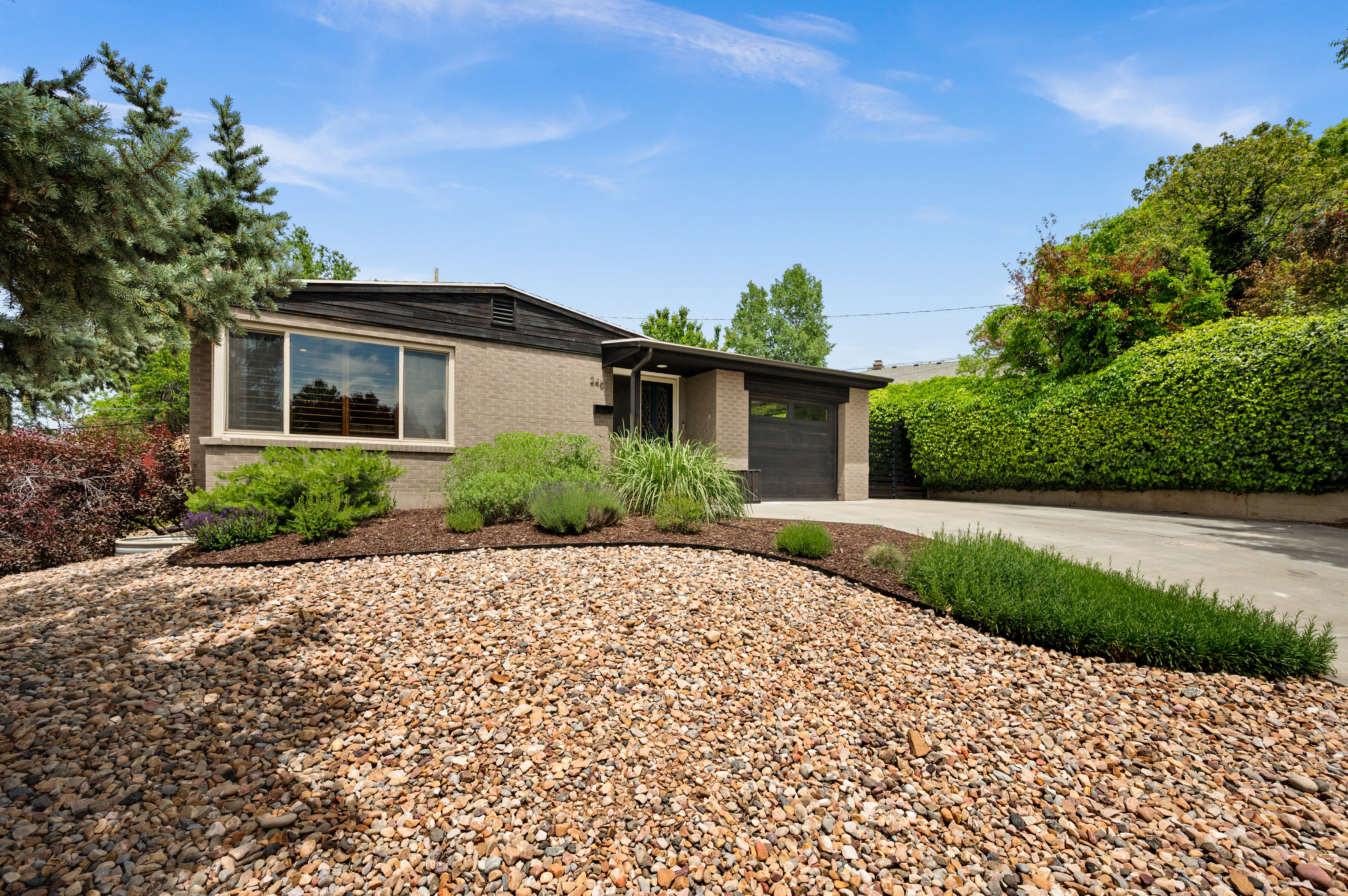 Brick exterior featuring a dark garage door, large front window with plantation shutters, low-maintenance gravel landscaping, and mature shrubbery
