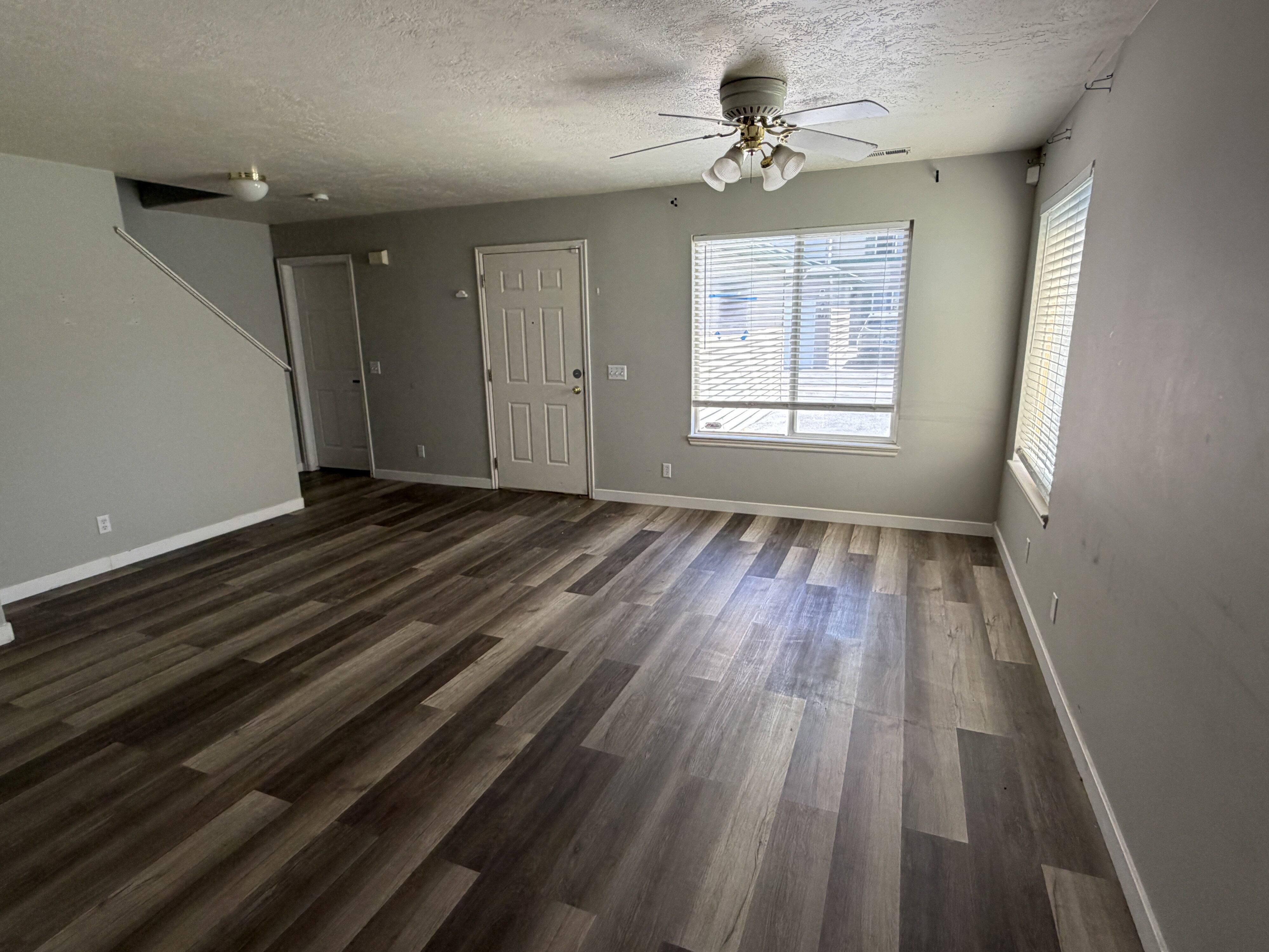 Spacious living area featuring wood-finish flooring, neutral wall tones, multiple windows with blinds, a ceiling fan with integrated lighting, and a paneled entry door
