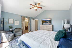 Vaulted ceiling bedroom featuring a ceiling fan, a doorway with drapes leading to a bathroom, and wood-finish flooring visible in the bathroom