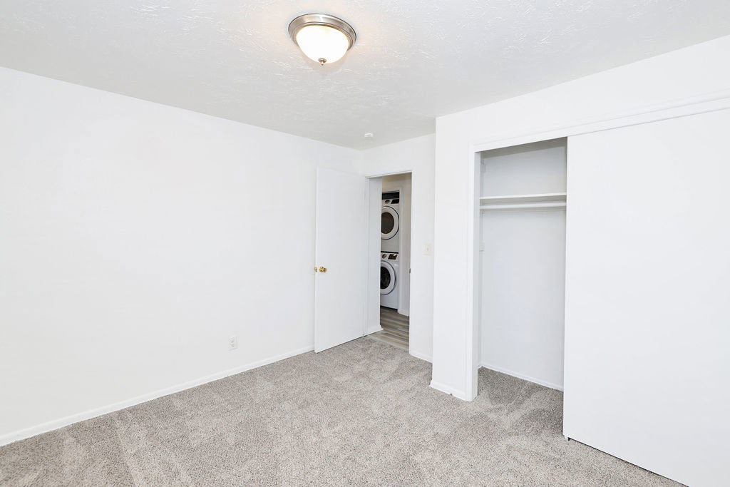 Carpeted room featuring a ceiling-mounted light fixture, sliding closet doors, and a laundry closet with stacked appliances