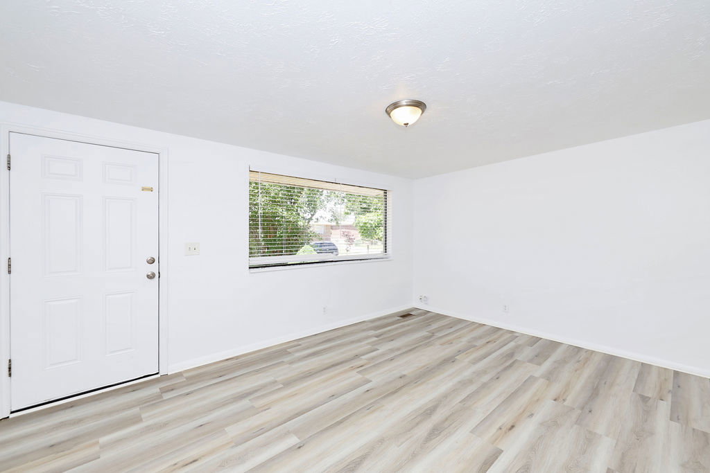 Bright interior space featuring a window with horizontal blinds, a white paneled entry door, wood-finish flooring, and a flush-mount ceiling light fixture